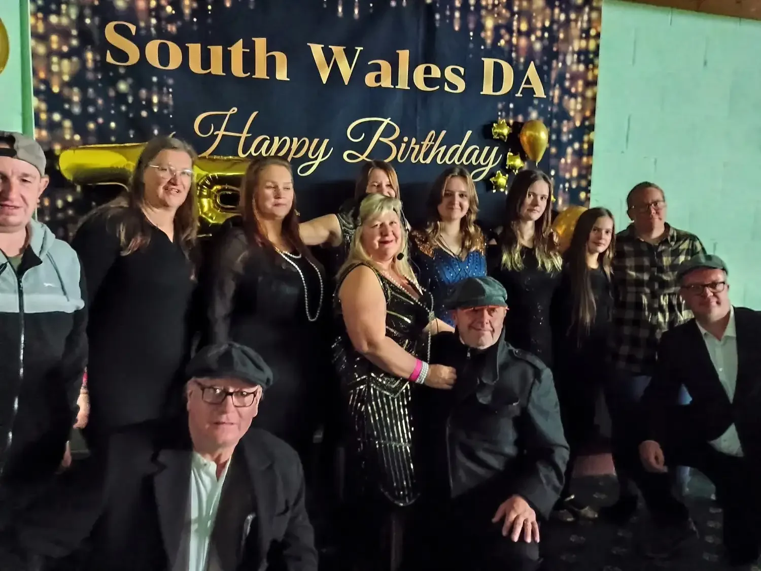 A group of ten people, mostly women, posing together at a birthday celebration with a backdrop that reads 'South Wales DA Happy Birthday,' decorated with gold balloons and star-shaped decorations.
