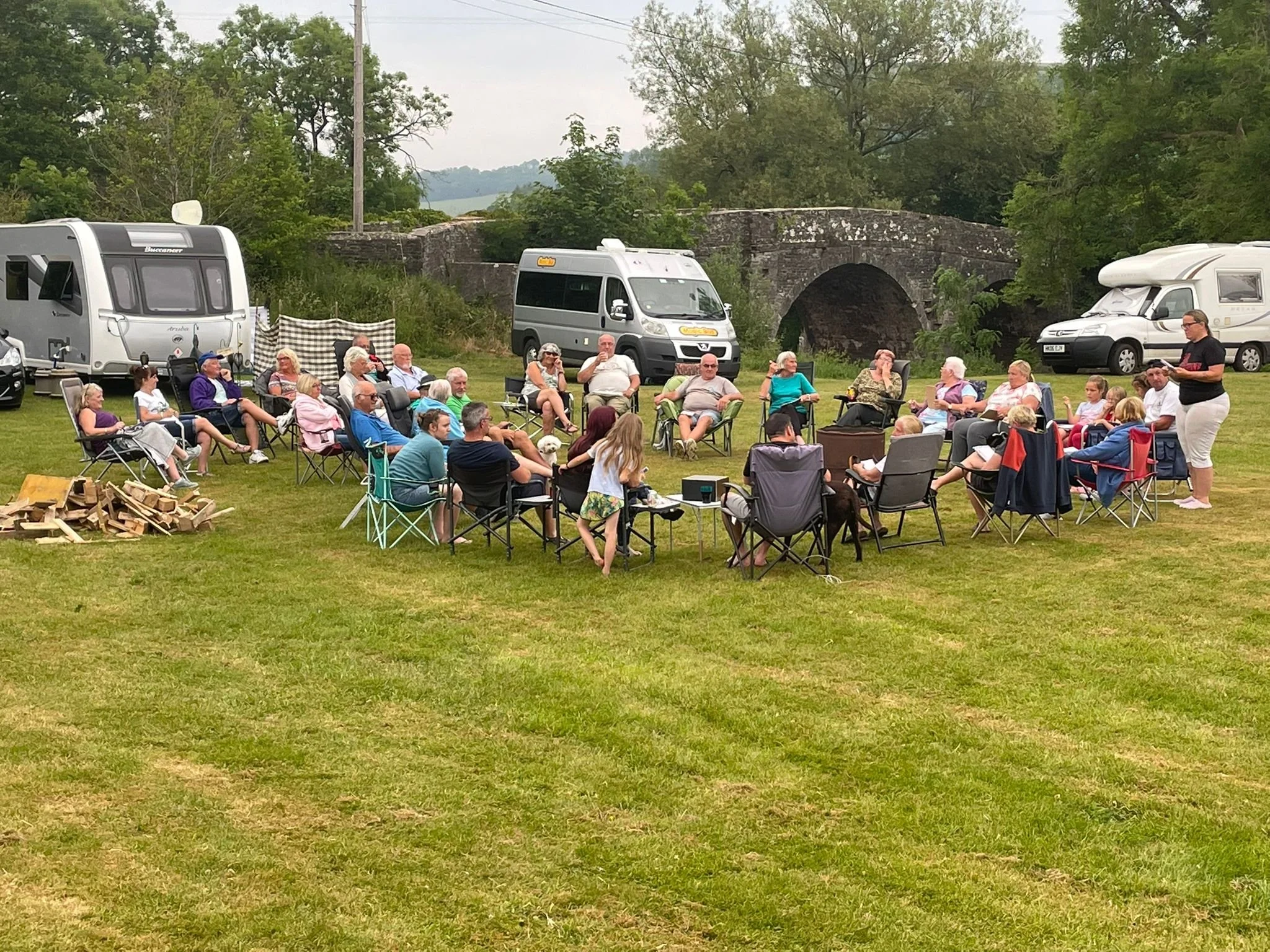 Group of people sitting in chairs outdoors on a grassy field, gathered around a speaker, with RVs and an old stone bridge in the background.