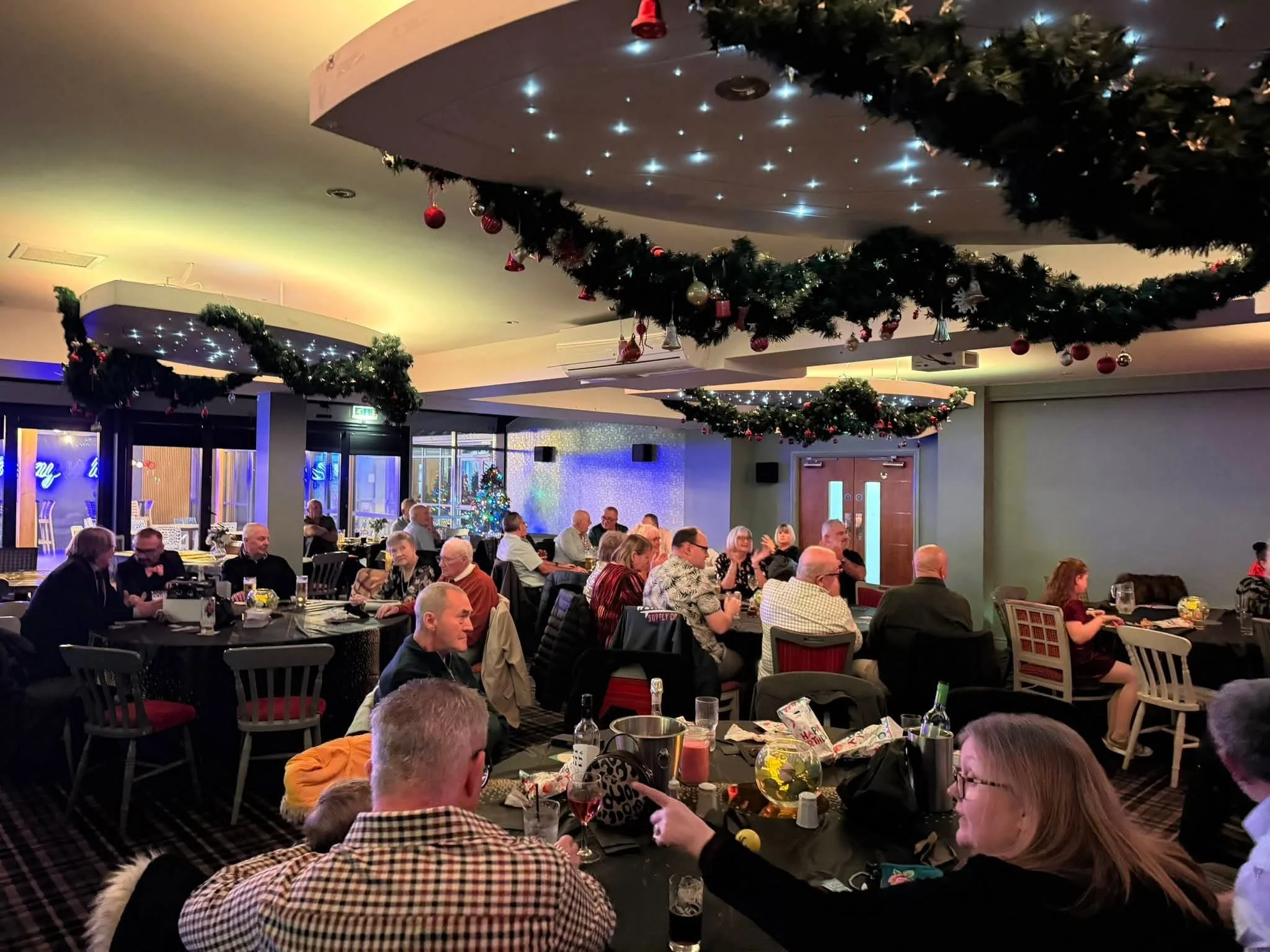 People gathered at tables in a decorated restaurant for a holiday celebration, with Christmas garlands and ornaments hanging from the ceiling.
