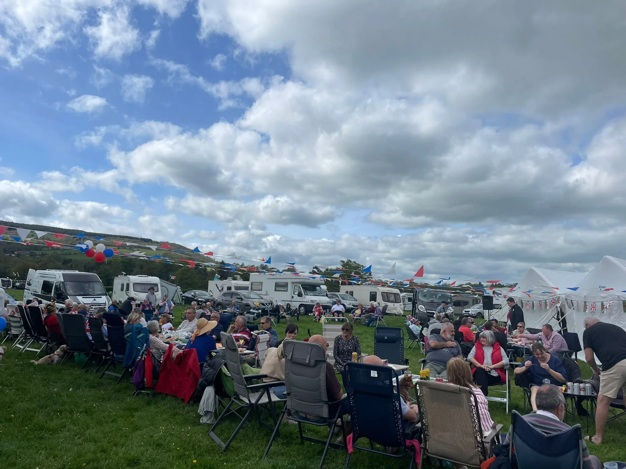 A large outdoor gathering with many people sitting on chairs at tables, celebrating on a grassy field decorated with red, white, and blue flags and balloons, with caravans and a cloudy sky in the background.