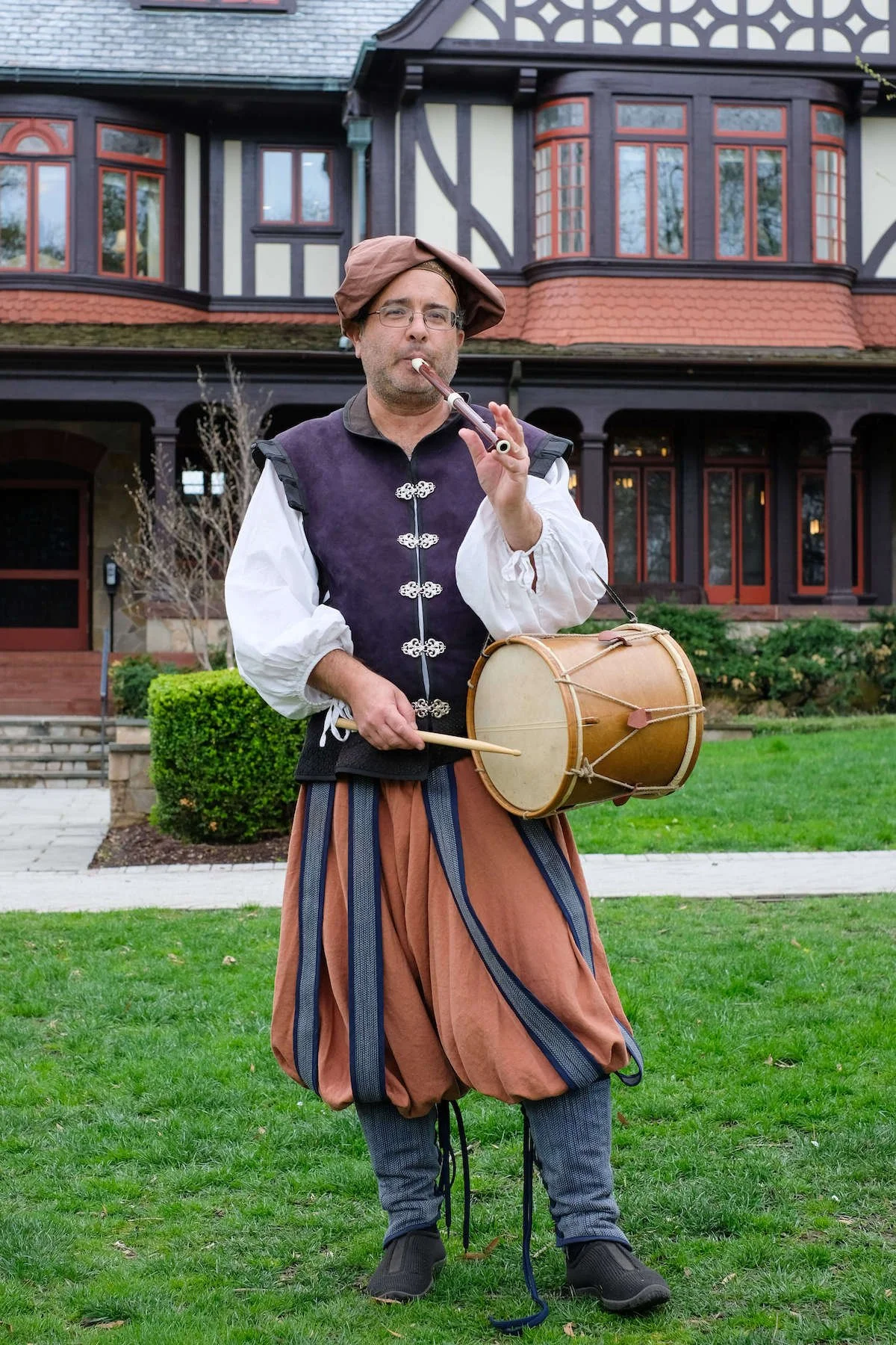 Man in Renaissance costume plays pipe and drum outside a historical building.