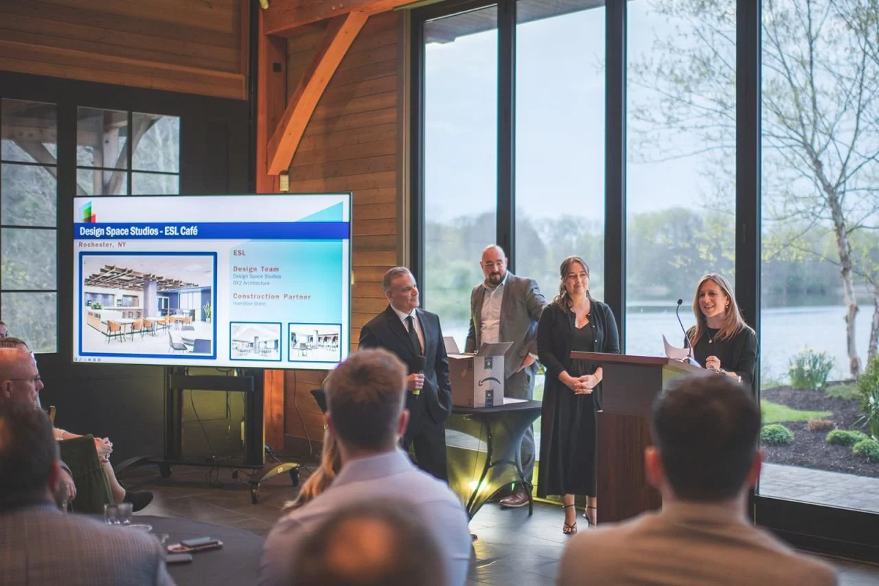 A group of four people presenting at a conference in a room with large windows overlooking a lake and trees, with an audience seated in front. A large screen displays information about a design project for ESL Café.