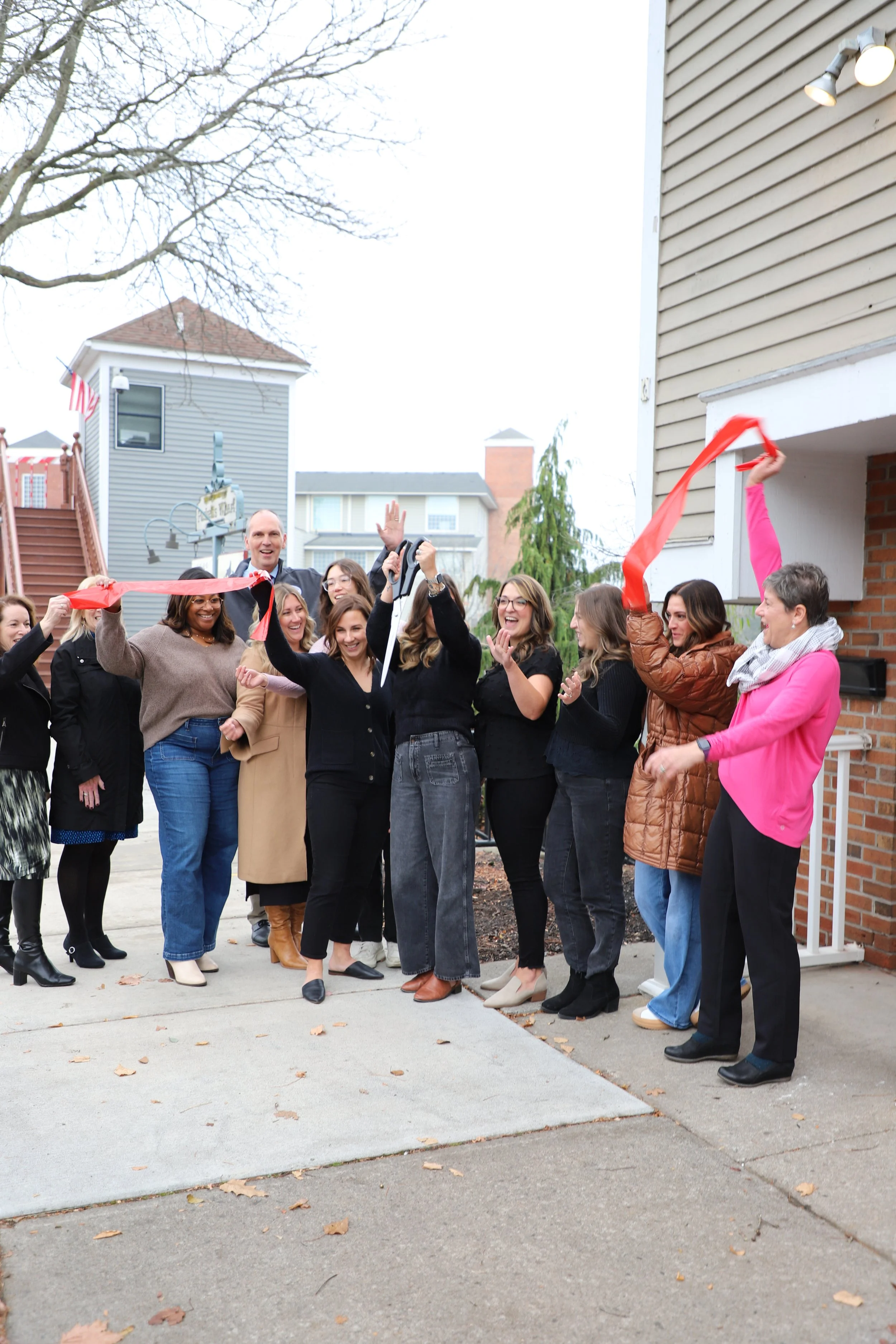 Design Space Studios team standing outside fairport office a ribbon-cutting event, some holding red ribbon pieces, on a sidewalk in front of a house.