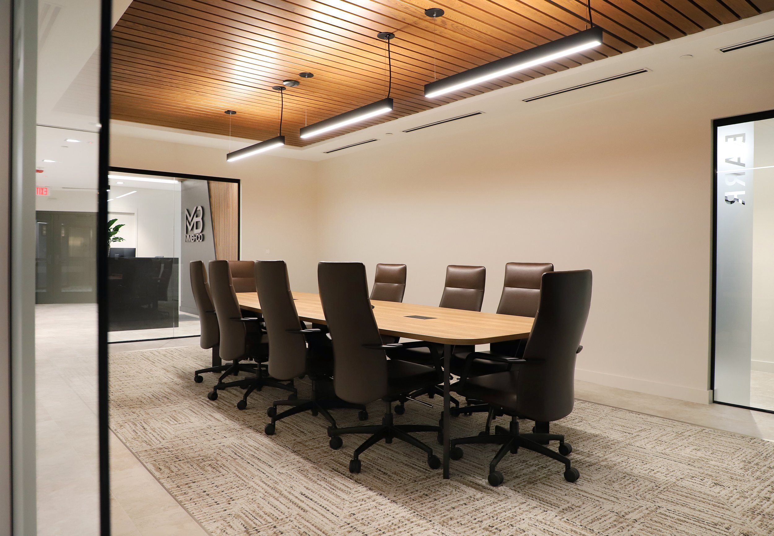 A modern conference room with a wooden ceiling, a long wooden table, and nine black leather office chairs on a beige rug. There are pendant lights hanging from the ceiling and glass walls with company logos.