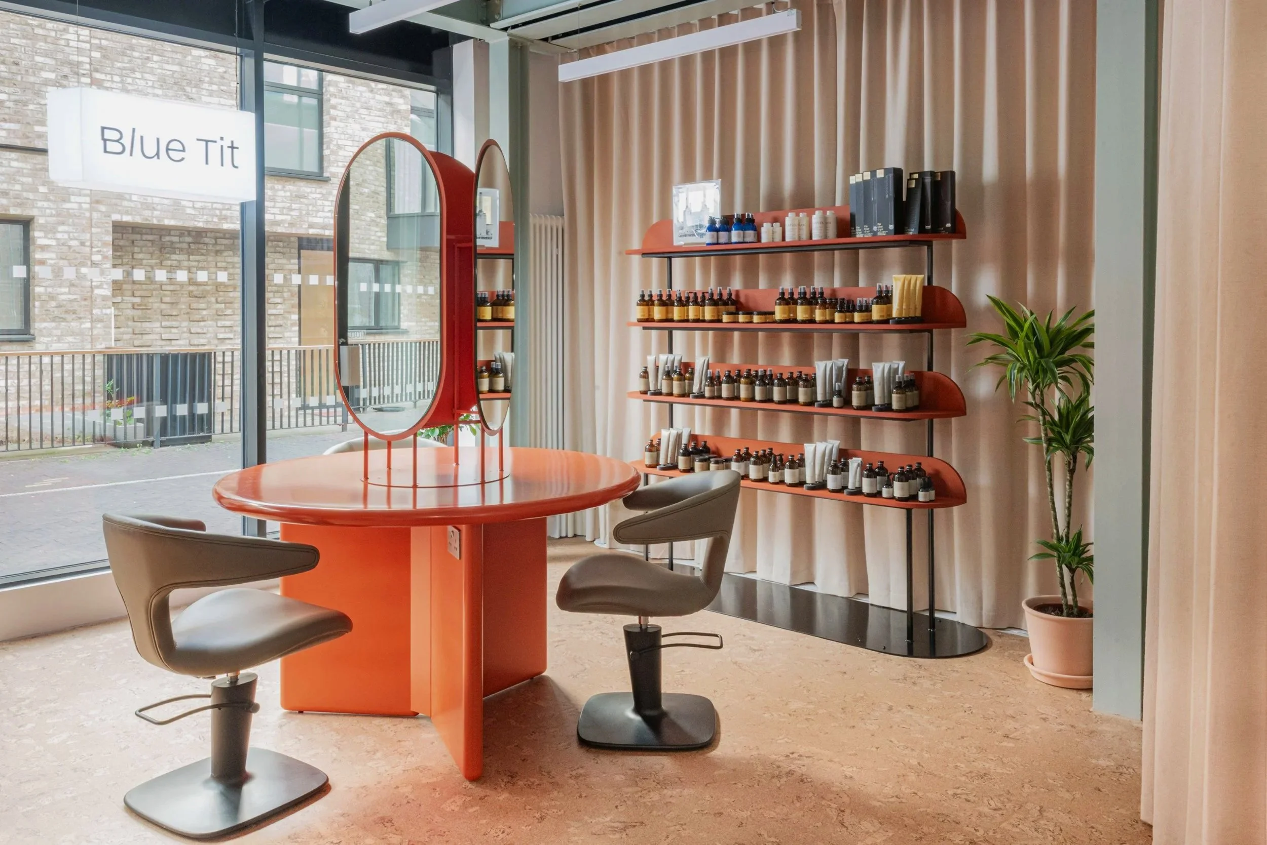 Interior of a beauty or skincare studio with a round orange table, gray chairs, shelves with bottles, and a potted plant near a large window with a sign that reads 'Blue Tit'.