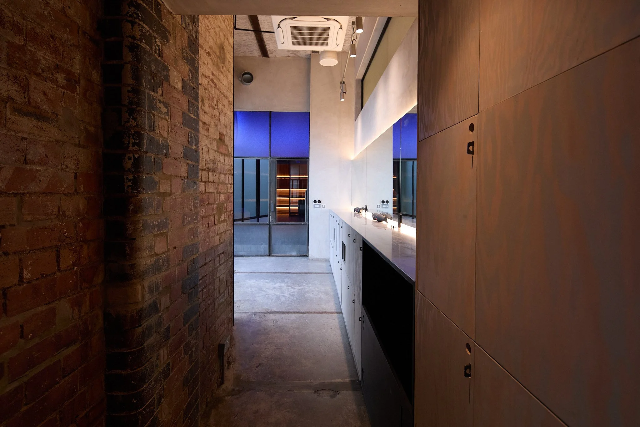 A modern kitchen with a concrete floor, exposed brick walls, white cabinetry, and a long white countertop. Large windows with blue lighting outside are visible in the background.