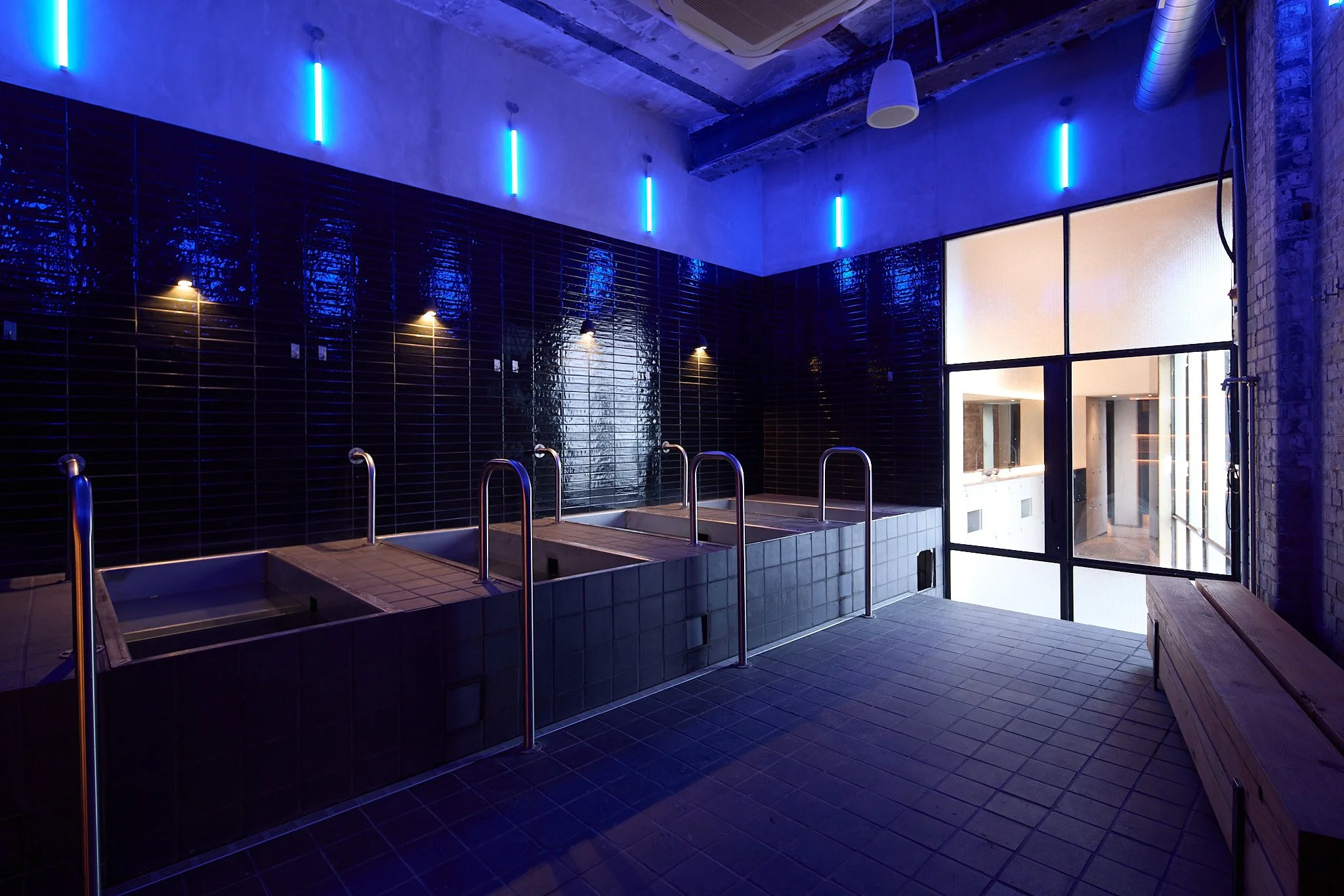 Indoor spa or bathing area with four rectangular beds, dark tiled walls, neon blue lighting, and a glass door leading outside.