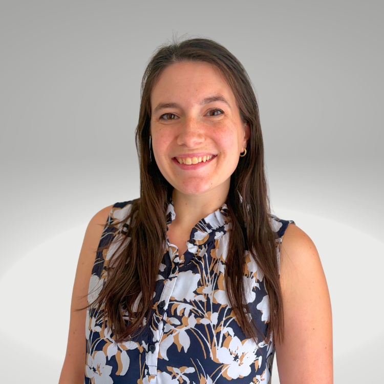 Young woman with long brown hair smiling, wearing a sleeveless floral blouse, standing against a gray background.