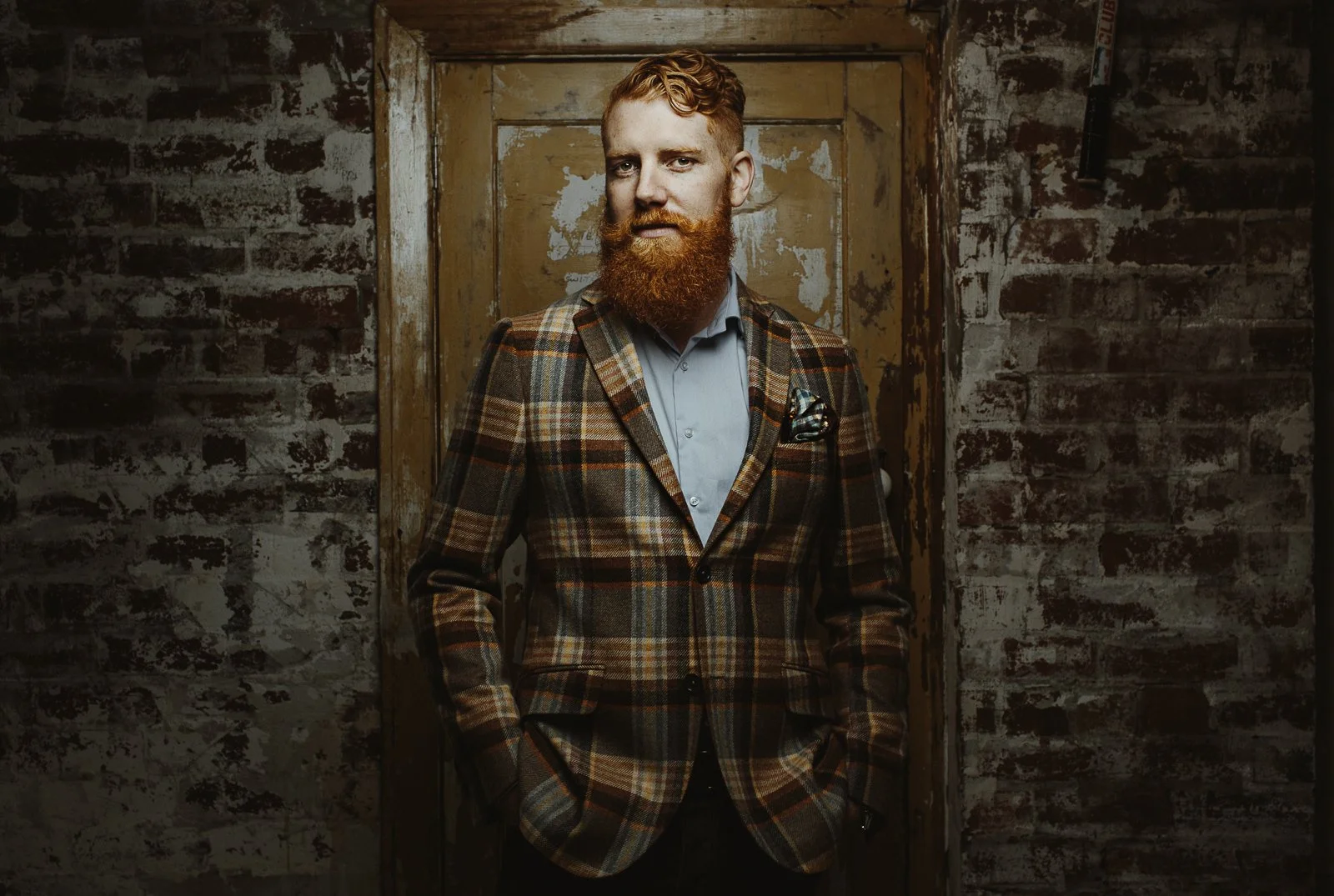 Man with red beard in plaid jacket standing against rustic brick wall.