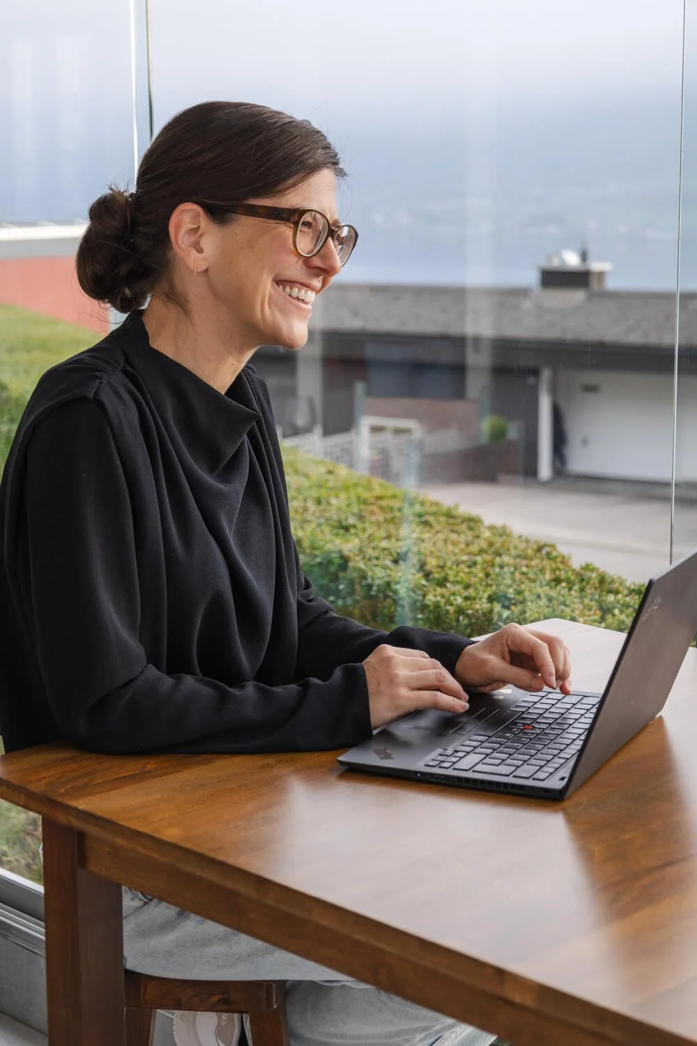 Yvonne smiling while typing on a laptop indoors with a view of greenery and buildings outside.