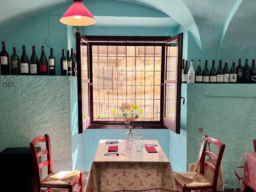 A cozy restaurant corner with a small wooden table set for two, with two glasses, a vase with flowers, and red napkins. There are two red wooden chairs, and a window with wooden shutters open, allowing sunlight to brighten the room. Bottles of wine line the ledge above the seating area, and a pink lampshade hangs from the ceiling.