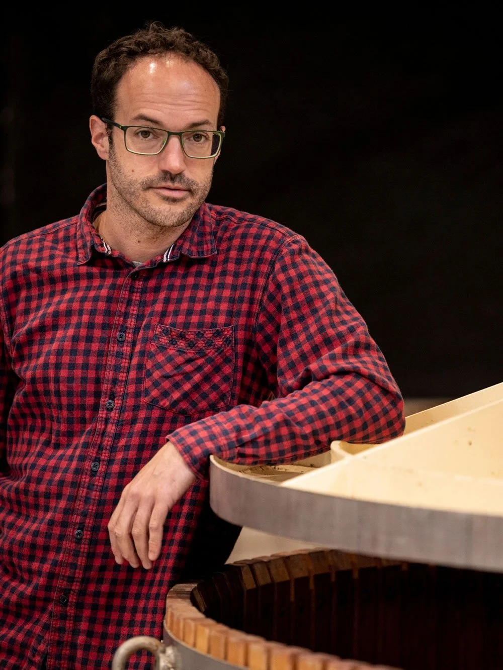 A man with glasses wearing a red and black checkered shirt, standing beside a large wooden vat, in front of a black background.