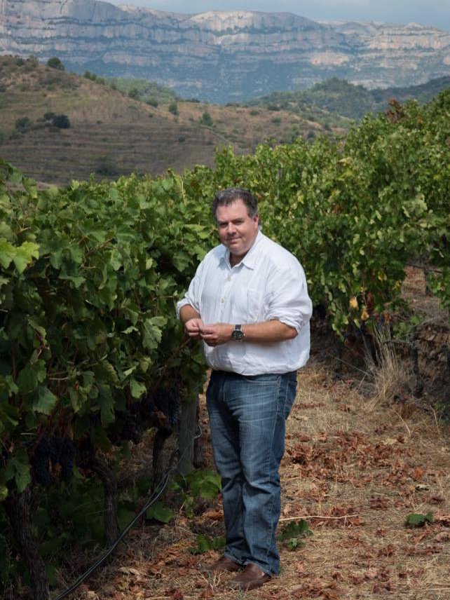 A man in a white shirt and jeans standing in a vineyard with green grapevines, with mountains and clear sky in the background.