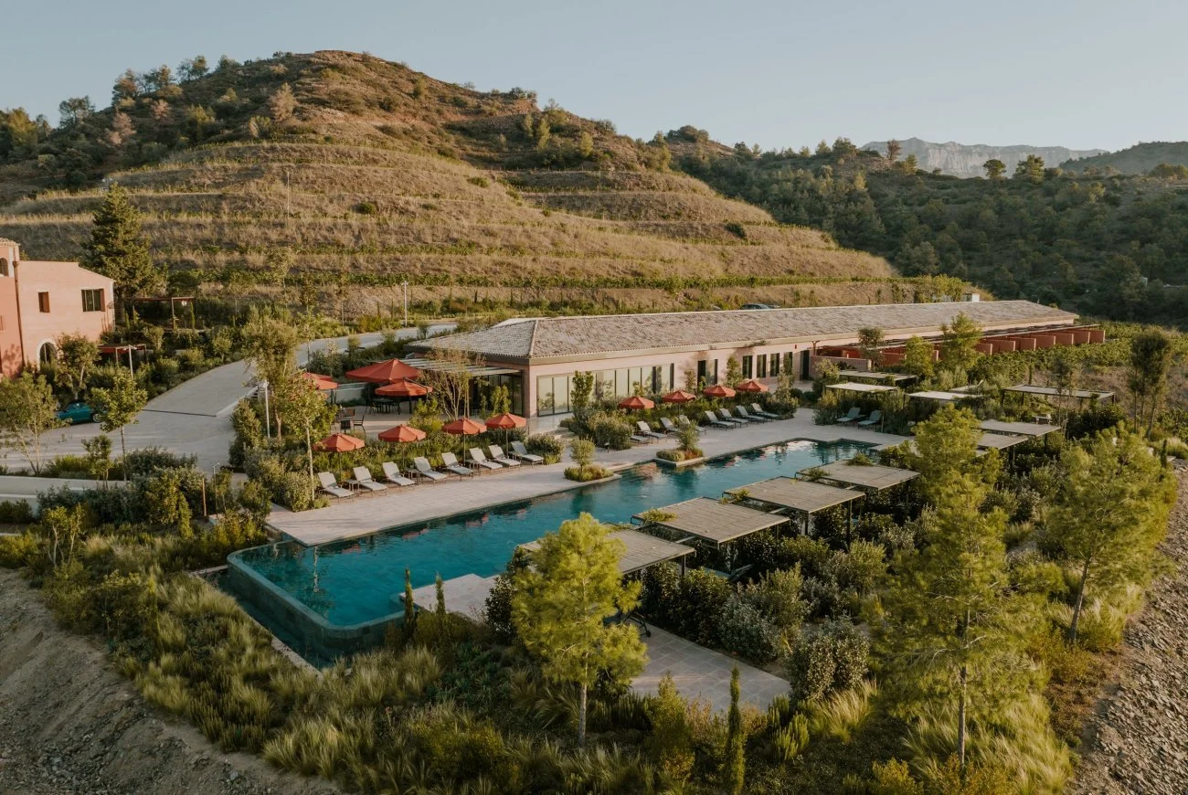 Aerial view of a luxury resort with a large swimming pool, surrounded by lounge chairs and umbrellas, nestled in a hilly, green landscape.