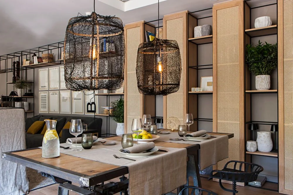 Modern dining room with a wooden table set with plates, glasses, and napkins, featuring two large woven hanging pendant lights. Background includes built-in shelves with decorative items and a cozy sitting area with cushions.