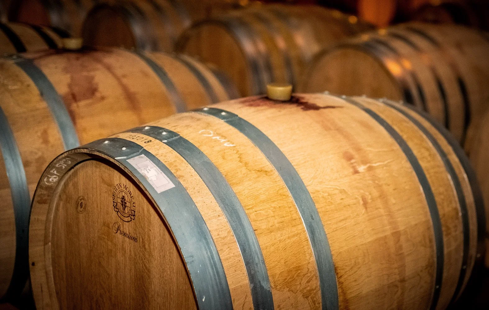 Row of wooden barrels in a cellar, with one barrel in the foreground marked with inscriptions and metal hoops.