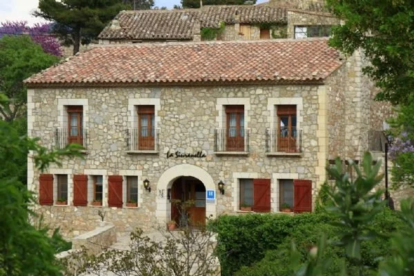 Stone house with red shutters and a tiled roof, surrounded by greenery.