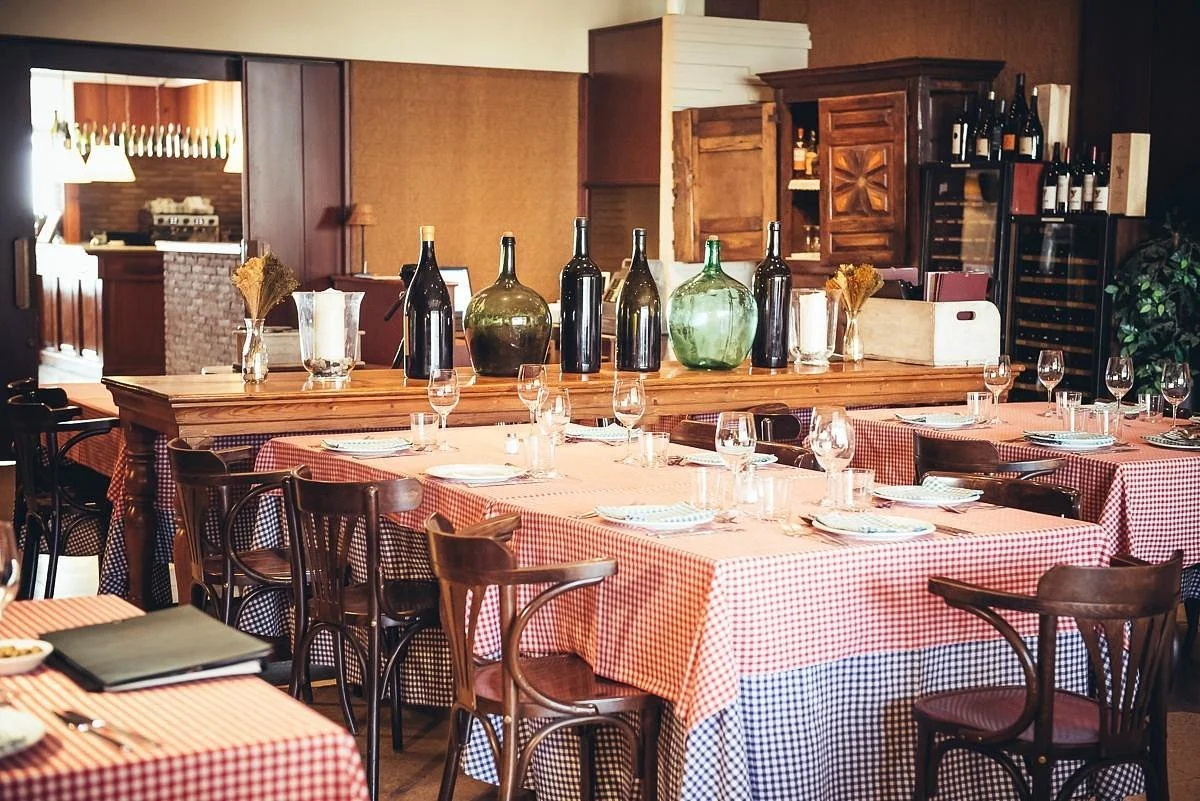 A restaurant dining area with tables covered in red and white checkered tablecloths, set with glasses, plates, and silverware. There are large wine bottles and vases with dried flowers on a central wooden table, and wooden chairs surrounding the tables. The background includes a wine cooler, shelves with bottles, and wooden cabinets.