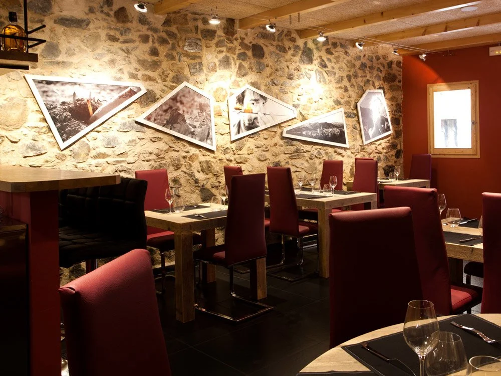 A cozy restaurant with a stone wall decorated with modern, angled black-and-white framed photographs, wooden tables set with wine glasses, and red upholstered chairs in warm lighting.