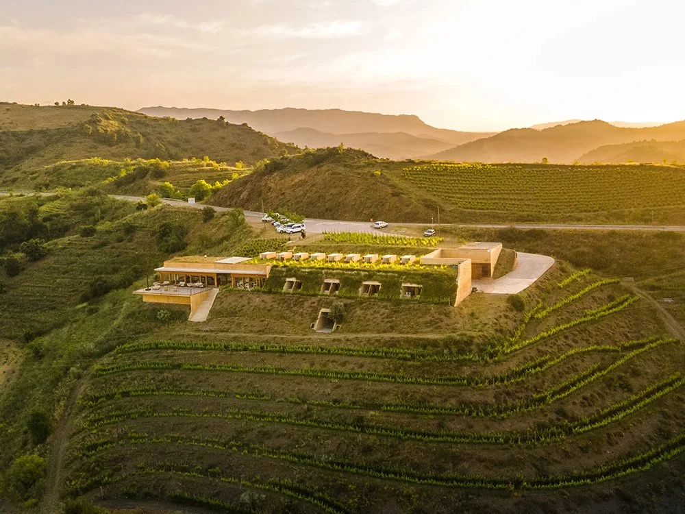 A modern house situated on a hillside with terraced vineyards, surrounded by rolling green hills and mountains at sunset.
