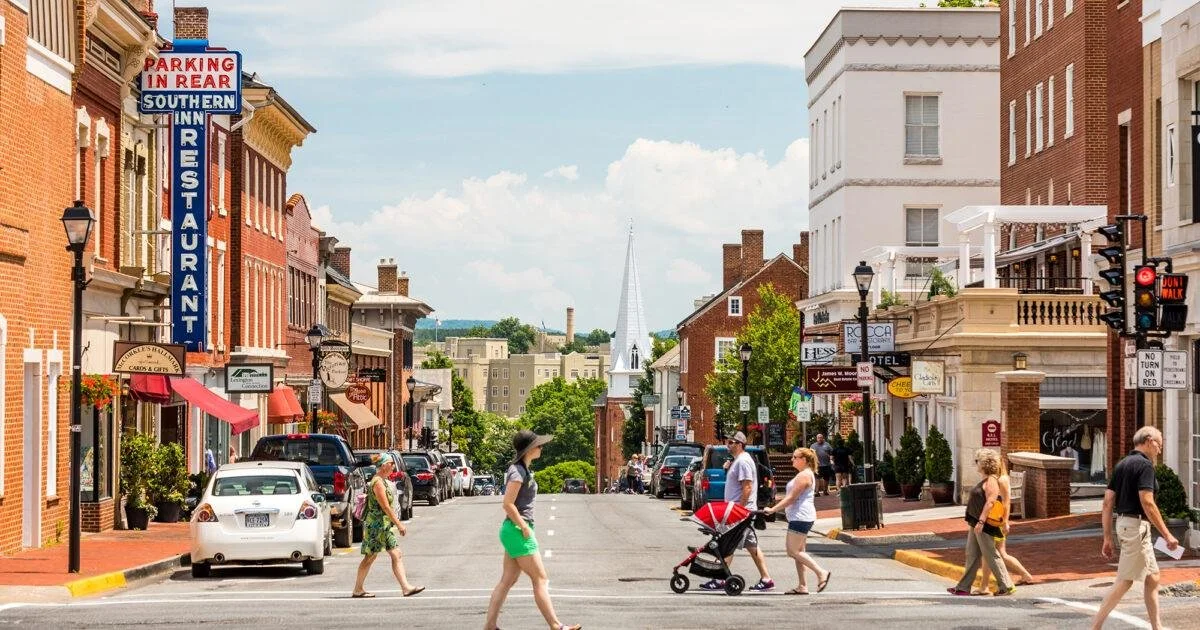 A lively small town street with people walking, shops, and parked cars. A tall church steeple is visible in the background under a partly cloudy sky.