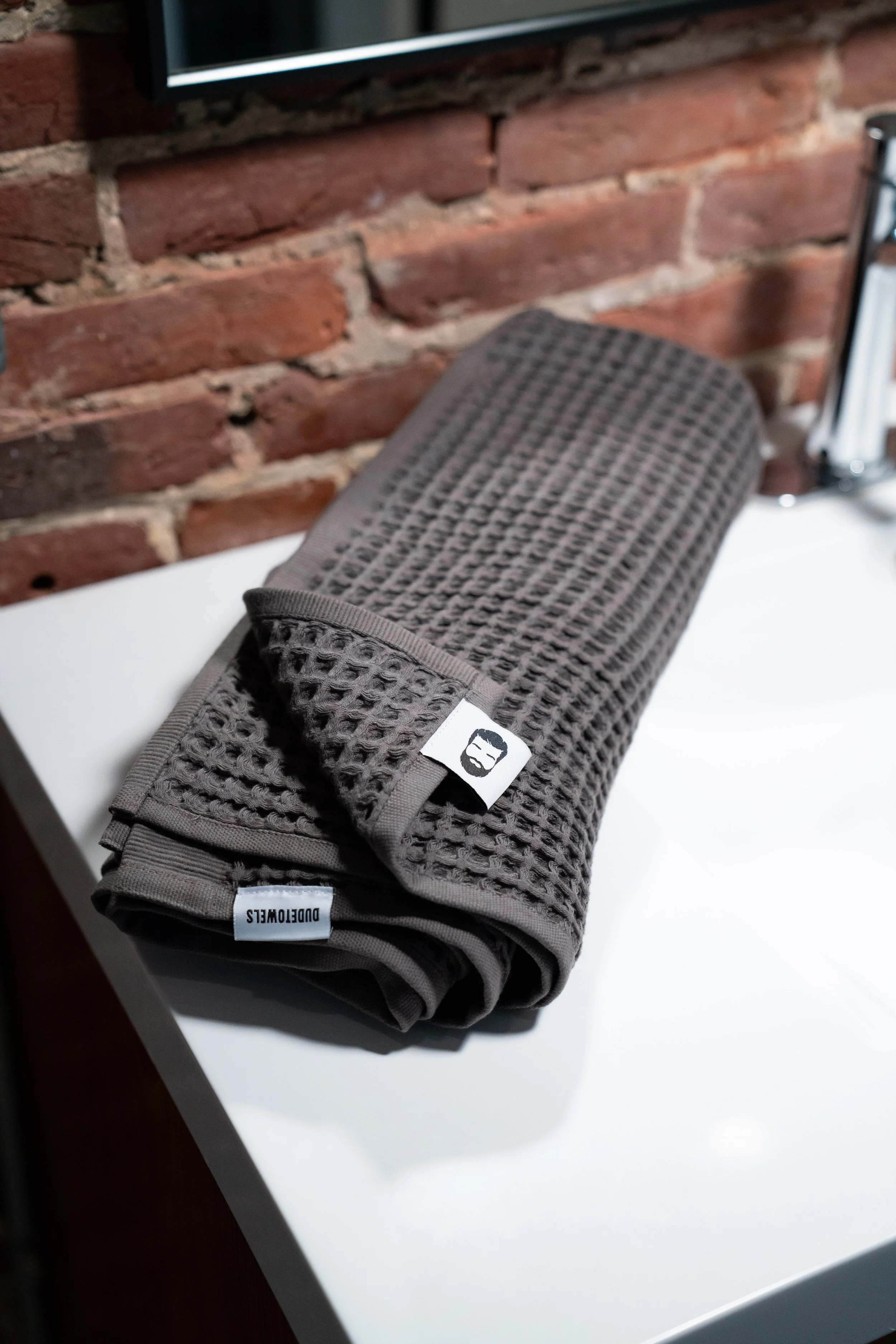 A folded dark gray waffle-patterned towel or cloth with tags that read 'DUDEWELS' and a logo featuring a bearded face with sunglasses, placed on a white surface near a brick wall.