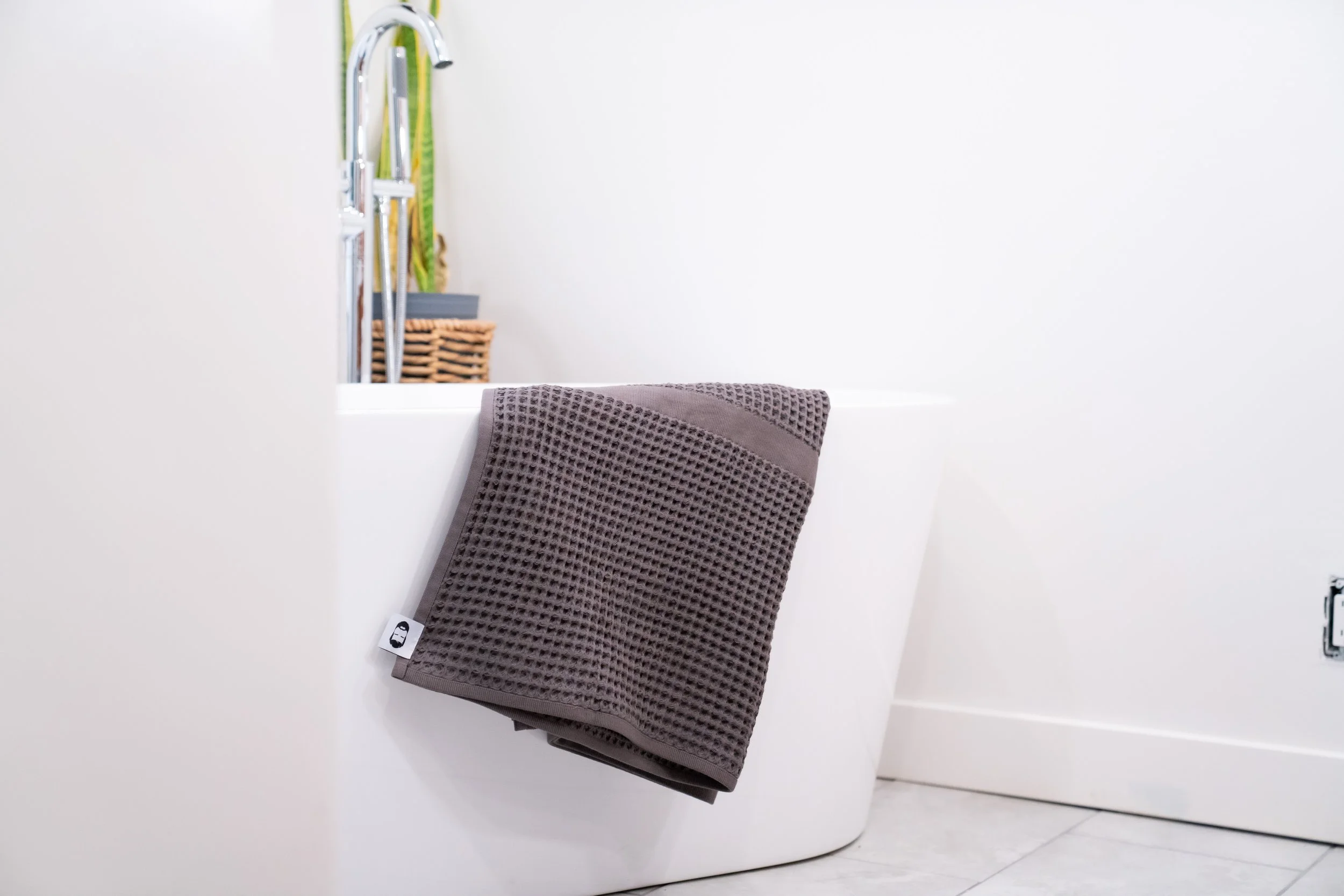 A white bathtub with a brown textured towel hanging on the edge, a chrome faucet, and a wicker basket with towels in the background.