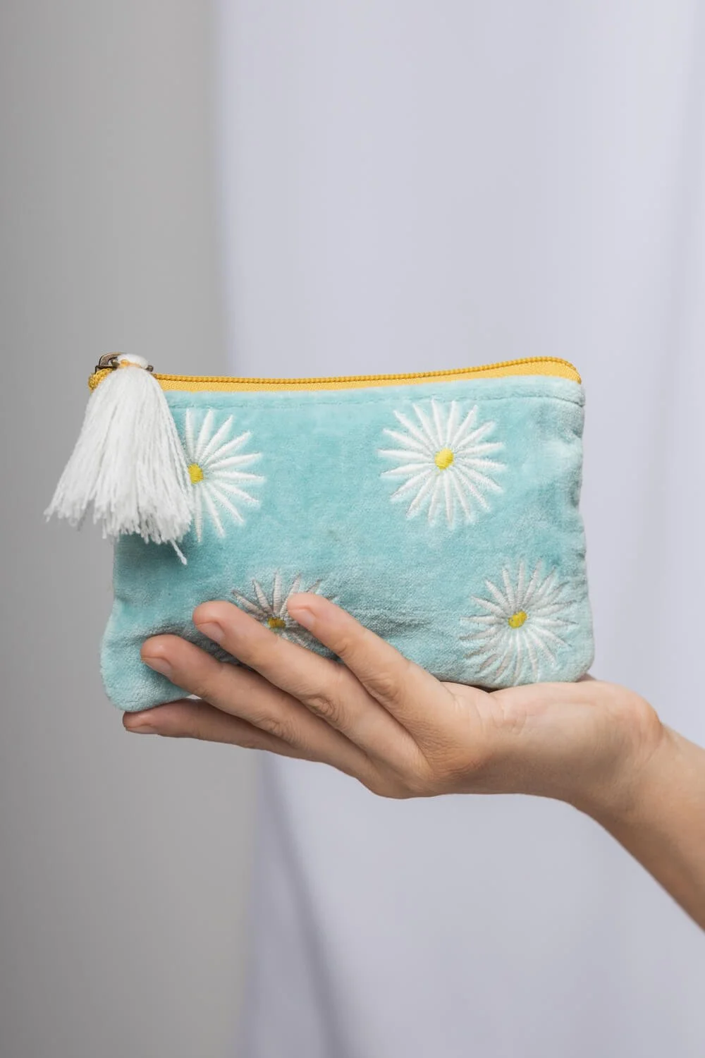 Turquoise pouch with white daisy embroidery and a yellow zipper, held by a hand, featuring a white tassel.
