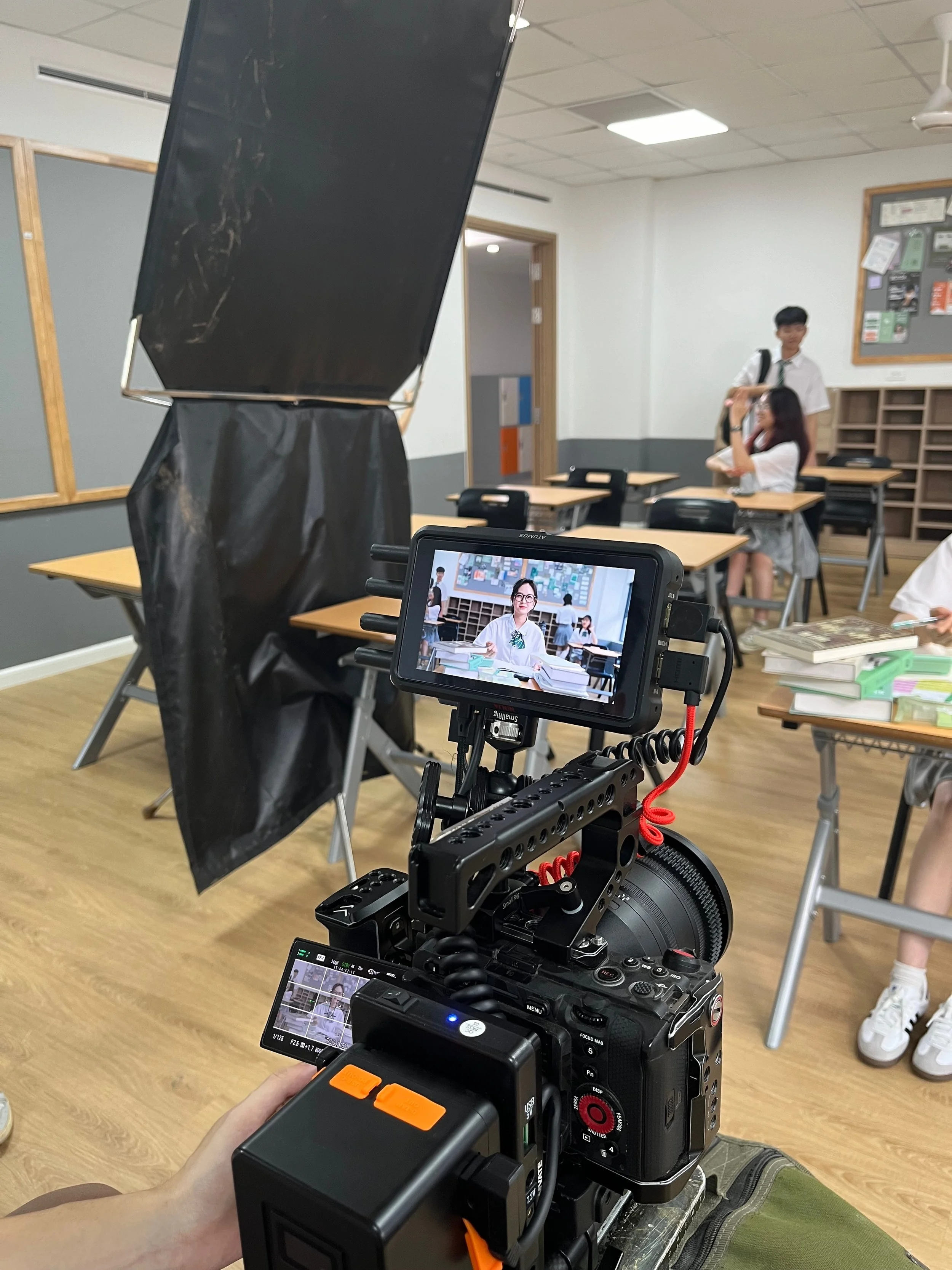 A camera on a tripod recording students in a classroom, with a monitor displaying the live feed. The classroom has students sitting at desks, including a girl adjusting her glasses and a boy standing nearby.
