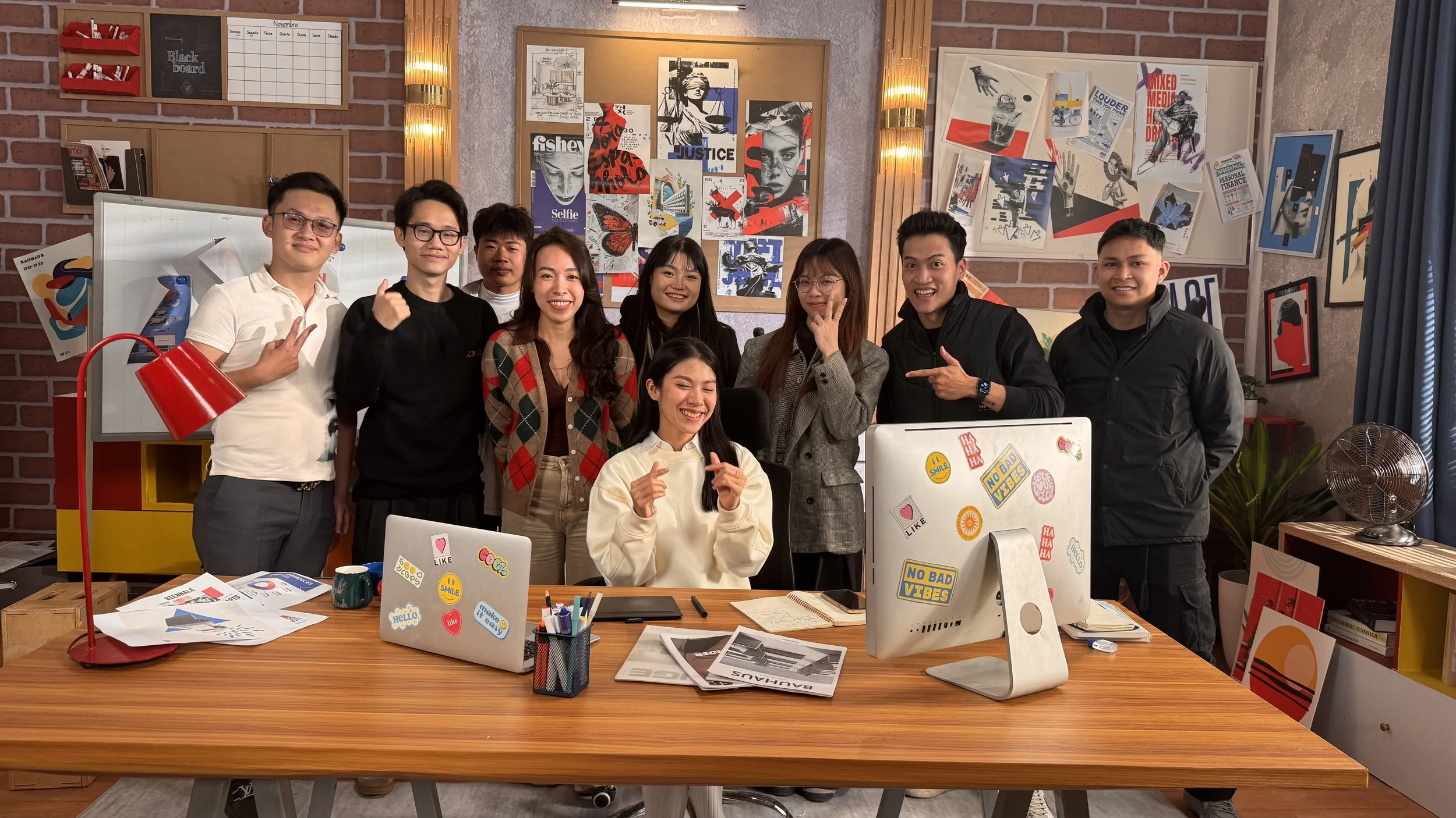 Group of nine people smiling and posing in an office decorated with colorful posters and artwork, with a desk in the foreground containing laptops, papers, and office supplies.