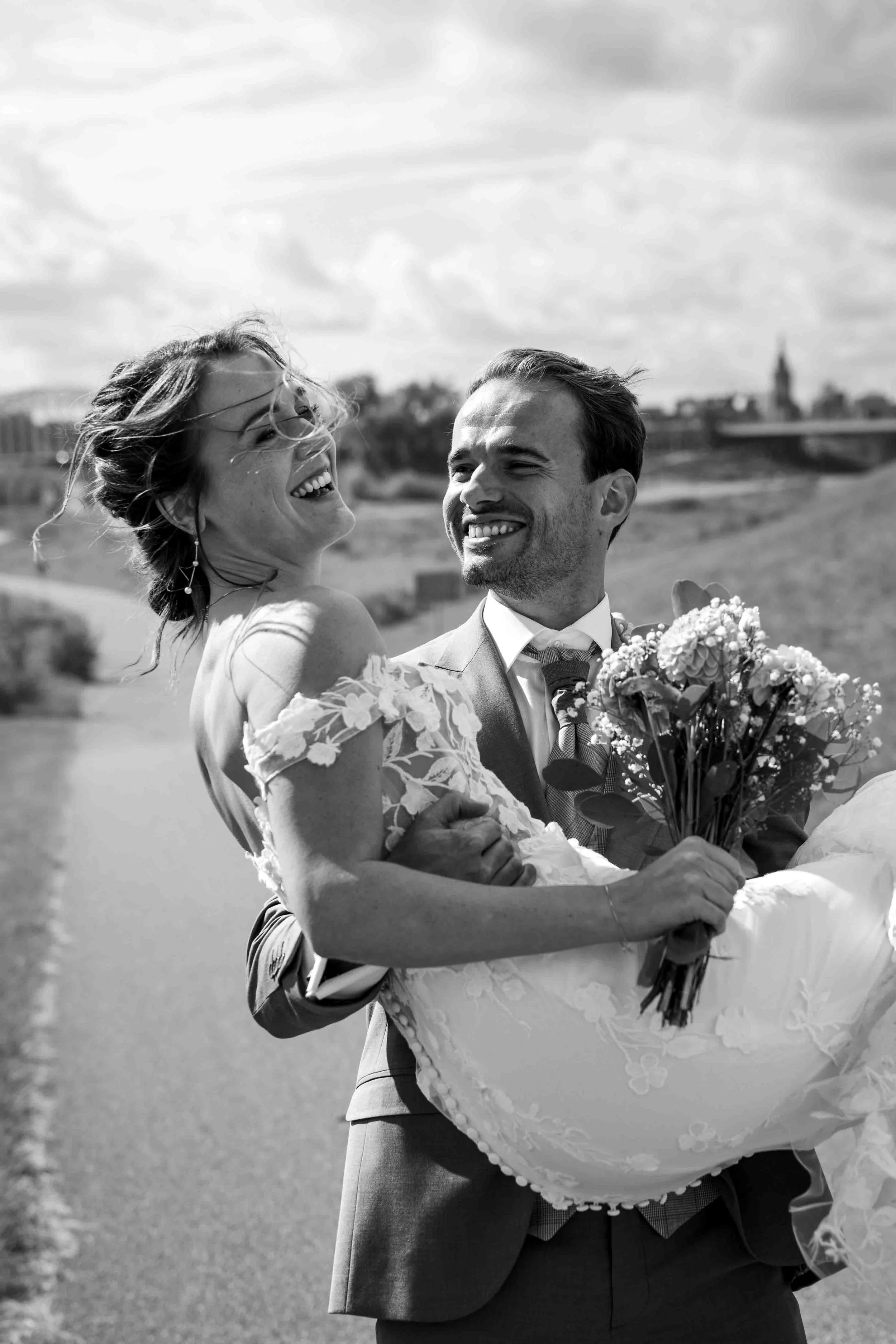 bruidegom tilt bruid op tijdens hun bruiloft aan de Waal in nijmegen, ze lachen allebei 