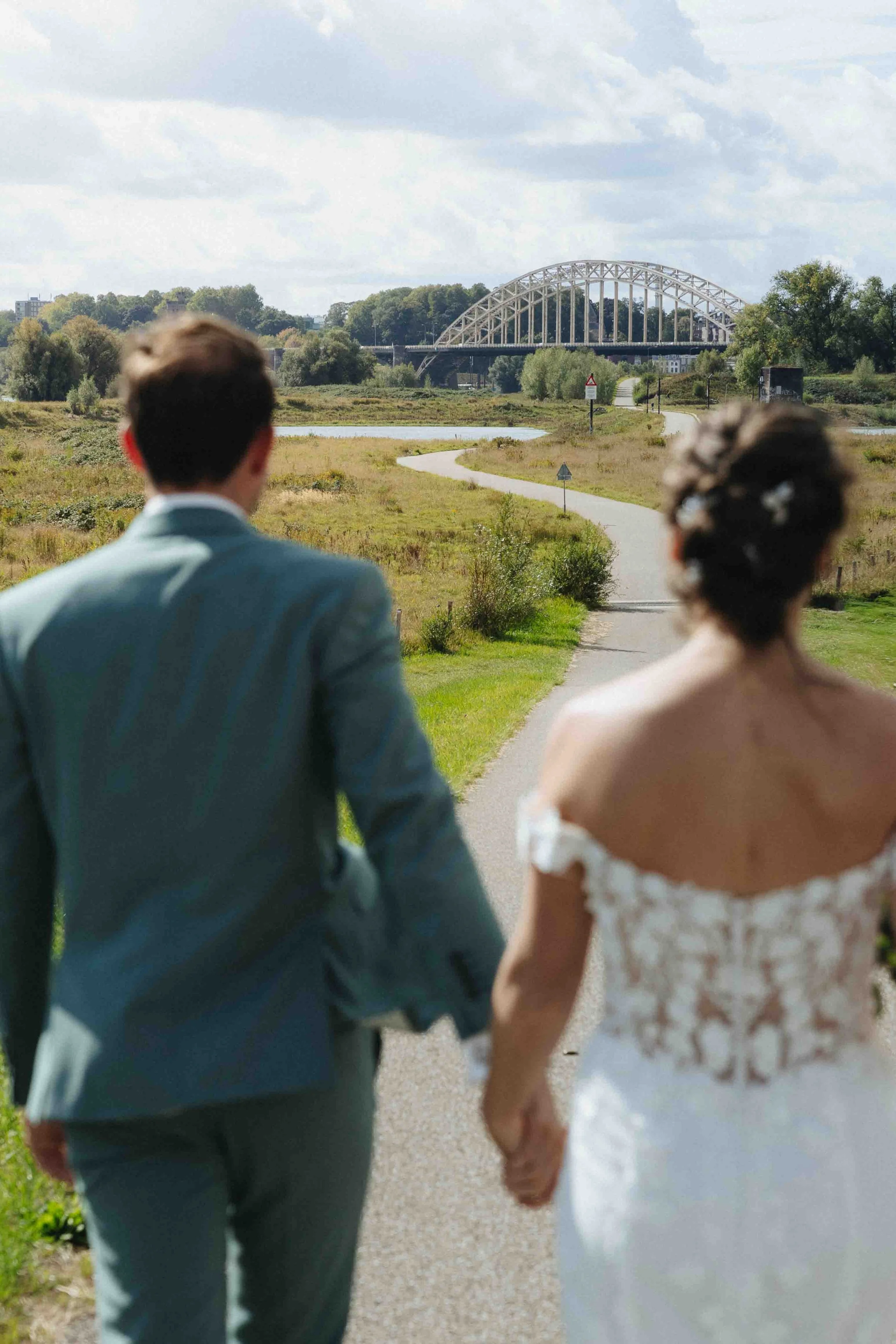 gelukkig bruidspaar in de zomer in nijmegen, wandelend langs de waal met de waalbrug op de achtergrond