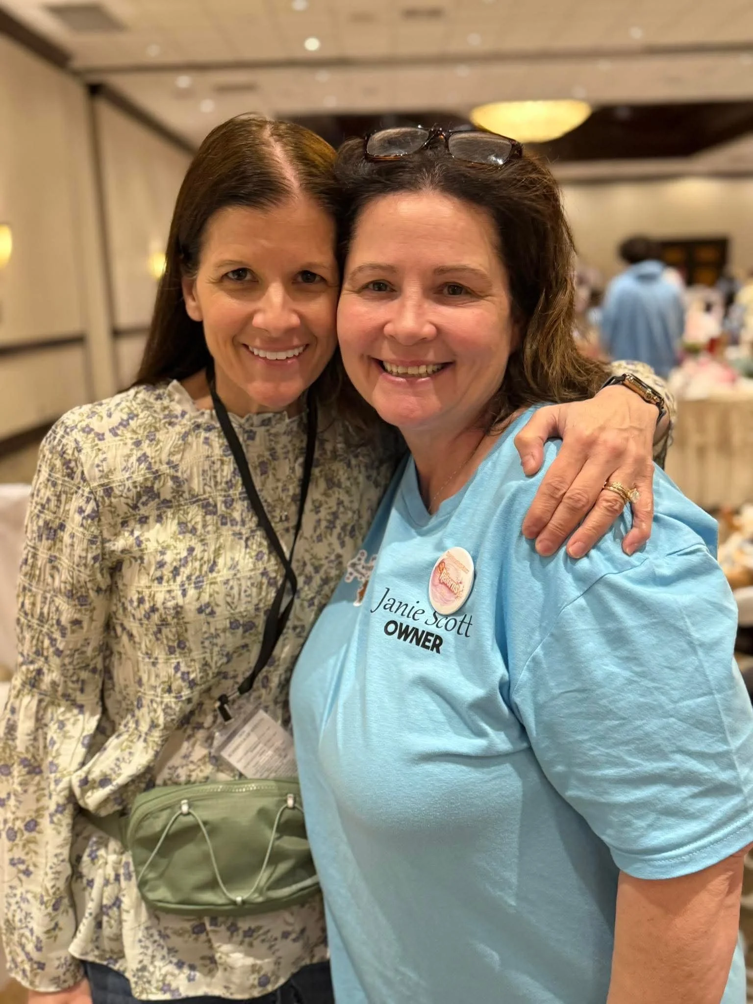 Two women smiling and hugging, in an indoor setting with blurred background of people and tables. One woman wears glasses on her head and a blue shirt with a name tag that reads "Janie Scott, OWNER". The other woman has dark hair and wears a patterned blouse.