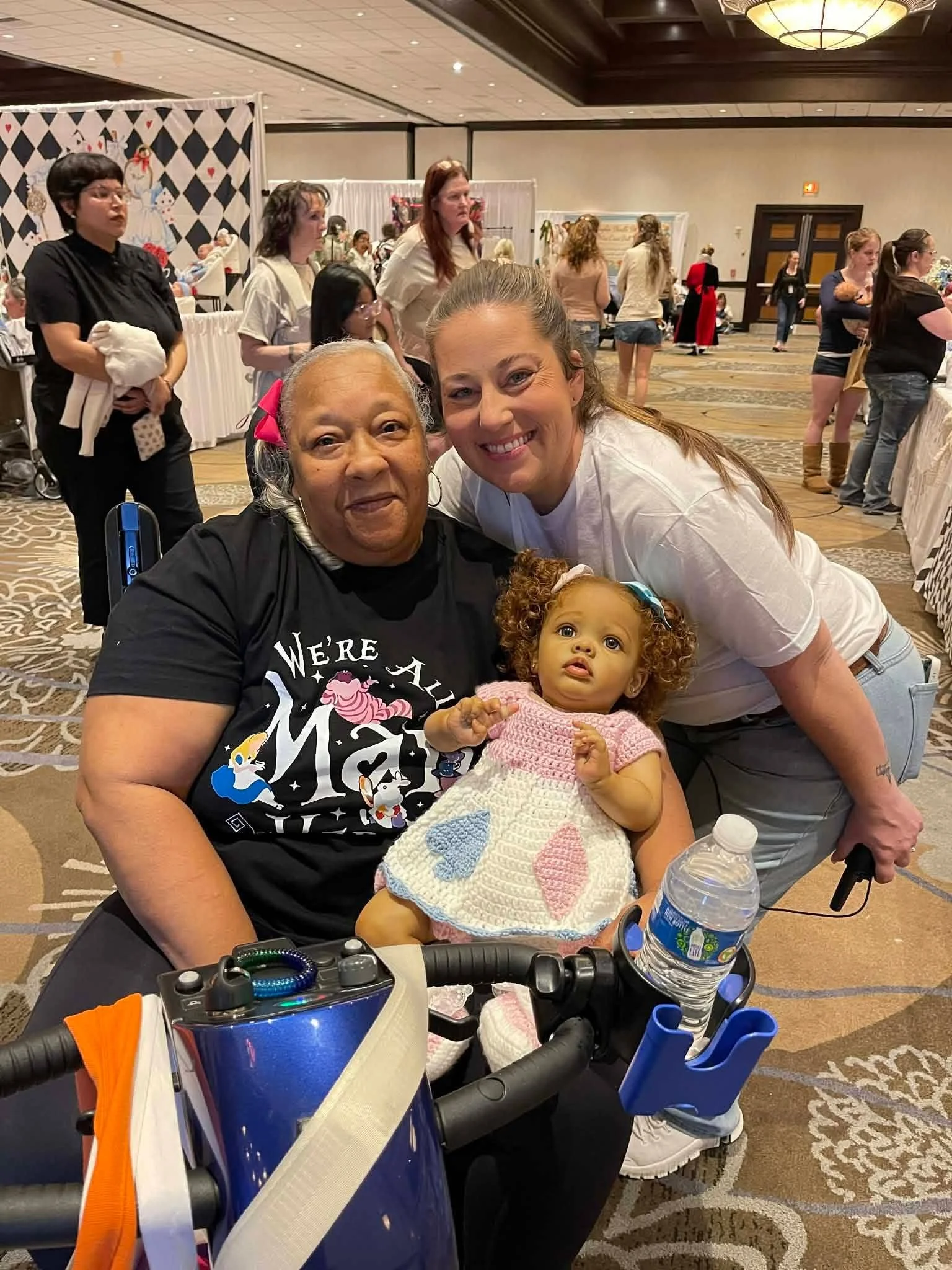 A group of people at a social event, with two women and a young girl in the foreground, smiling and posing for a photo. The background shows other attendees, booths or displays, and decorative elements.