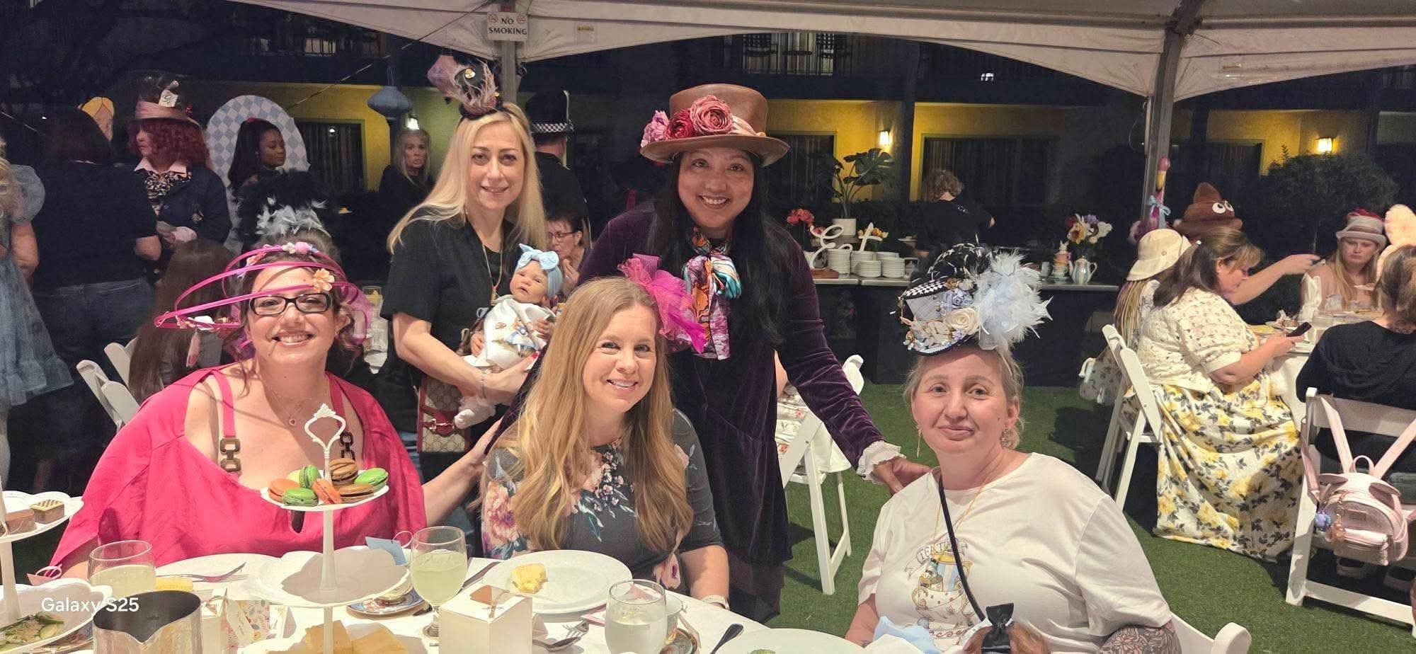 People celebrating at an outdoor party with women wearing elaborate hats, including a woman holding a baby, seated at a table with desserts and drinks, under a white tent at night.