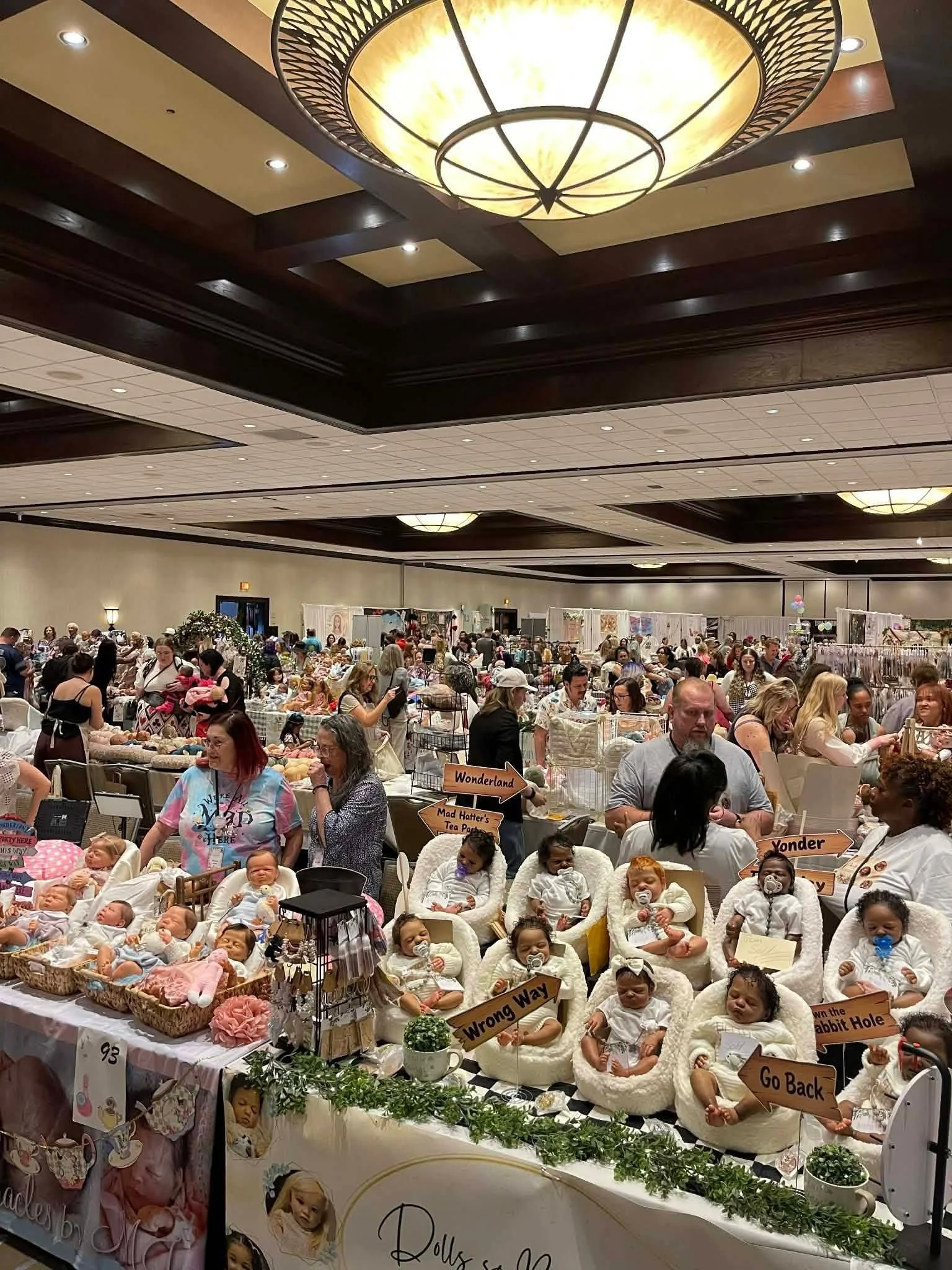 A crowded indoor event with multiple vendors selling dolls and stuffed toys. People are browsing and engaging at various tables, with signs like 'Wonderland,' 'Wrong Way,' and 'Go Back.'