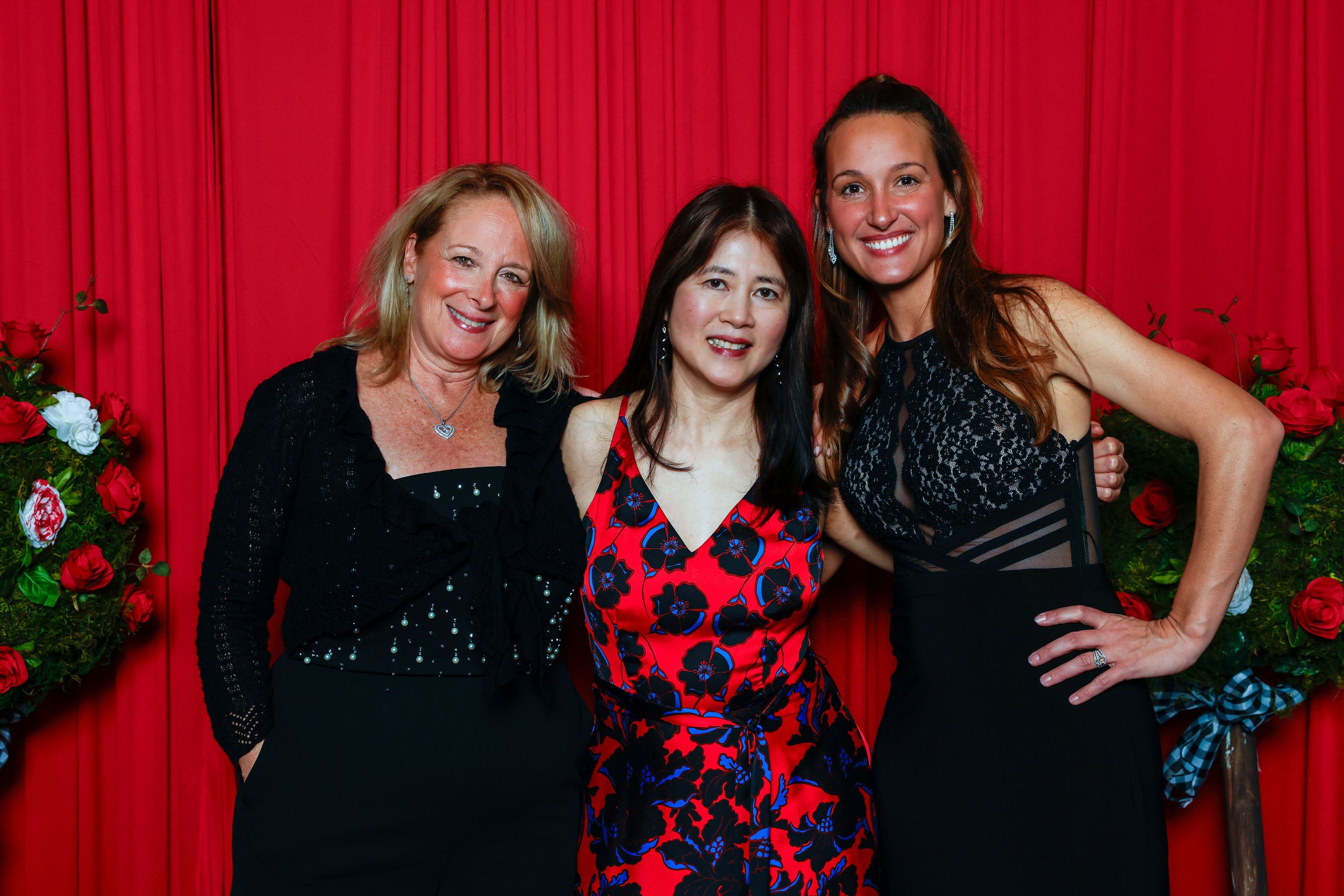 Three women standing in front of red curtains, smiling for the photo. One woman is wearing a black dress, another a red and black floral dress, and the third a black dress with sheer panels. There are red roses on either side of the women.