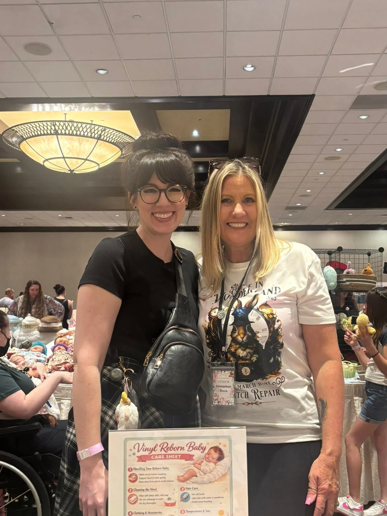 Two women smiling at a table at a craft fair. One woman wears glasses, has dark hair in a bun, and a black shirt. The other woman has blonde hair, is wearing a white t-shirt with a lion graphic, and has glasses on her head. There are children and oth