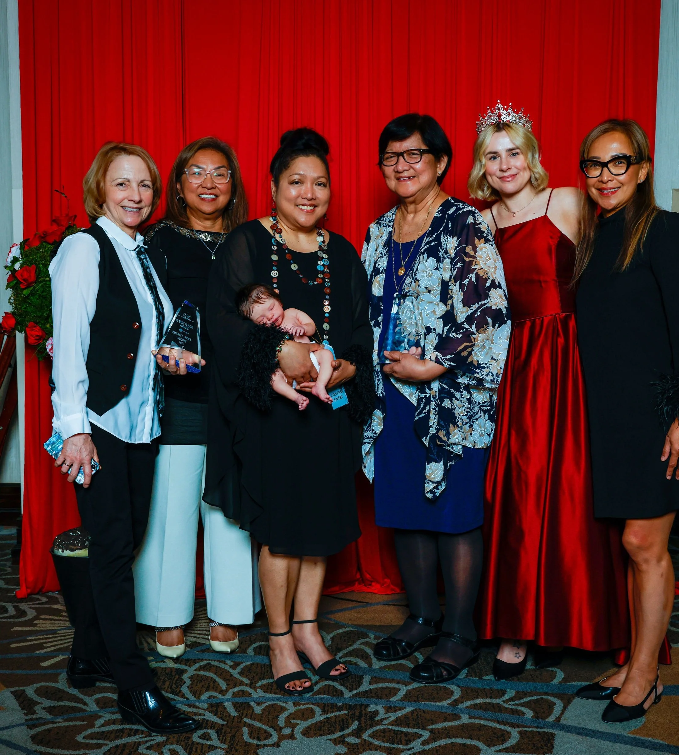 A group of seven women standing together in front of a red curtain at a celebration or awards event, with a woman in the center holding a small child. One woman is wearing a crown.