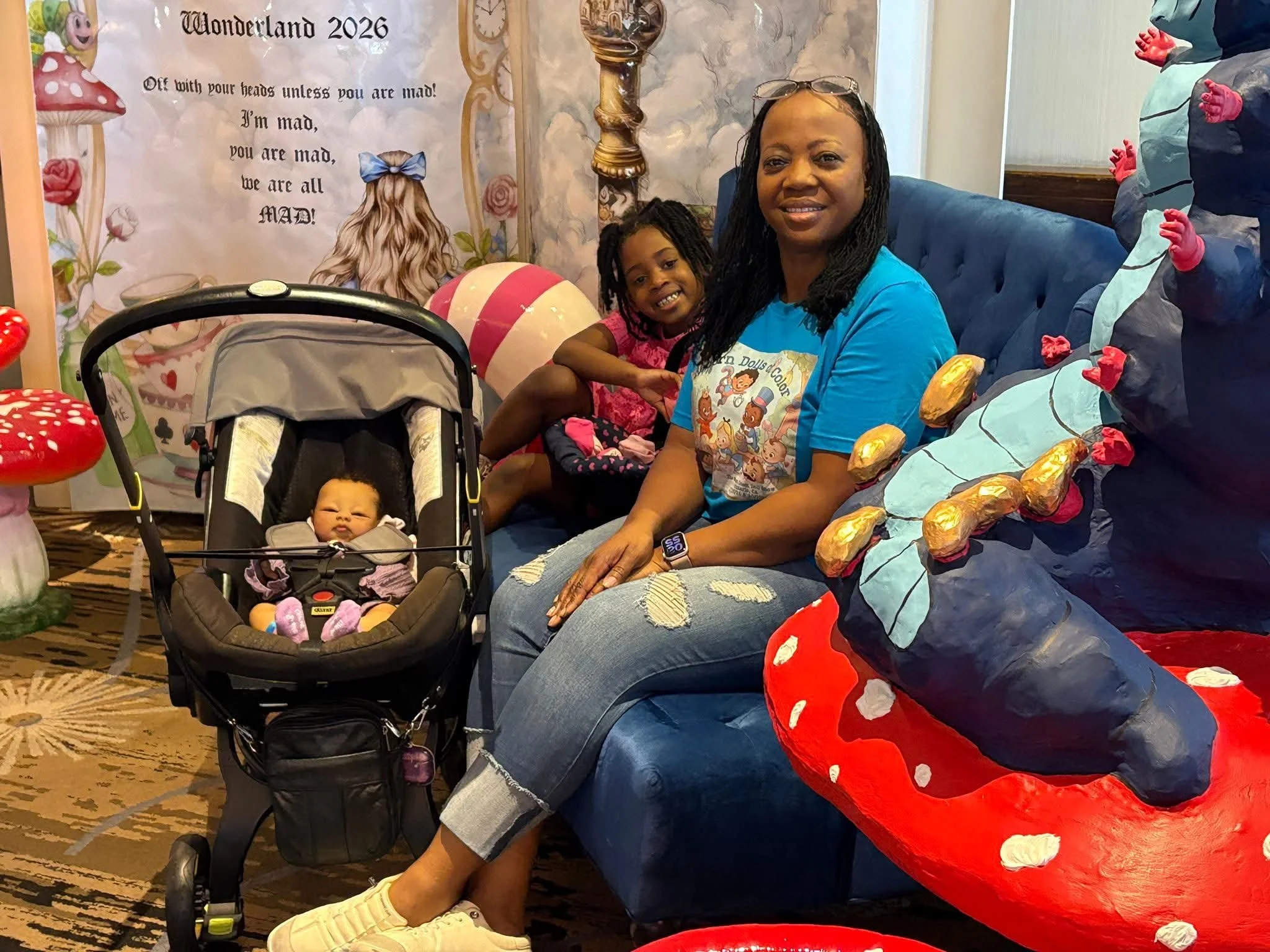 A smiling woman and a young girl sitting on a blue couch, with a baby in a car seat in front of them. There is a colorful backdrop with an Alice in Wonderland theme, including a banner that says "Wonderland 2026" and a quote from the story. The woman