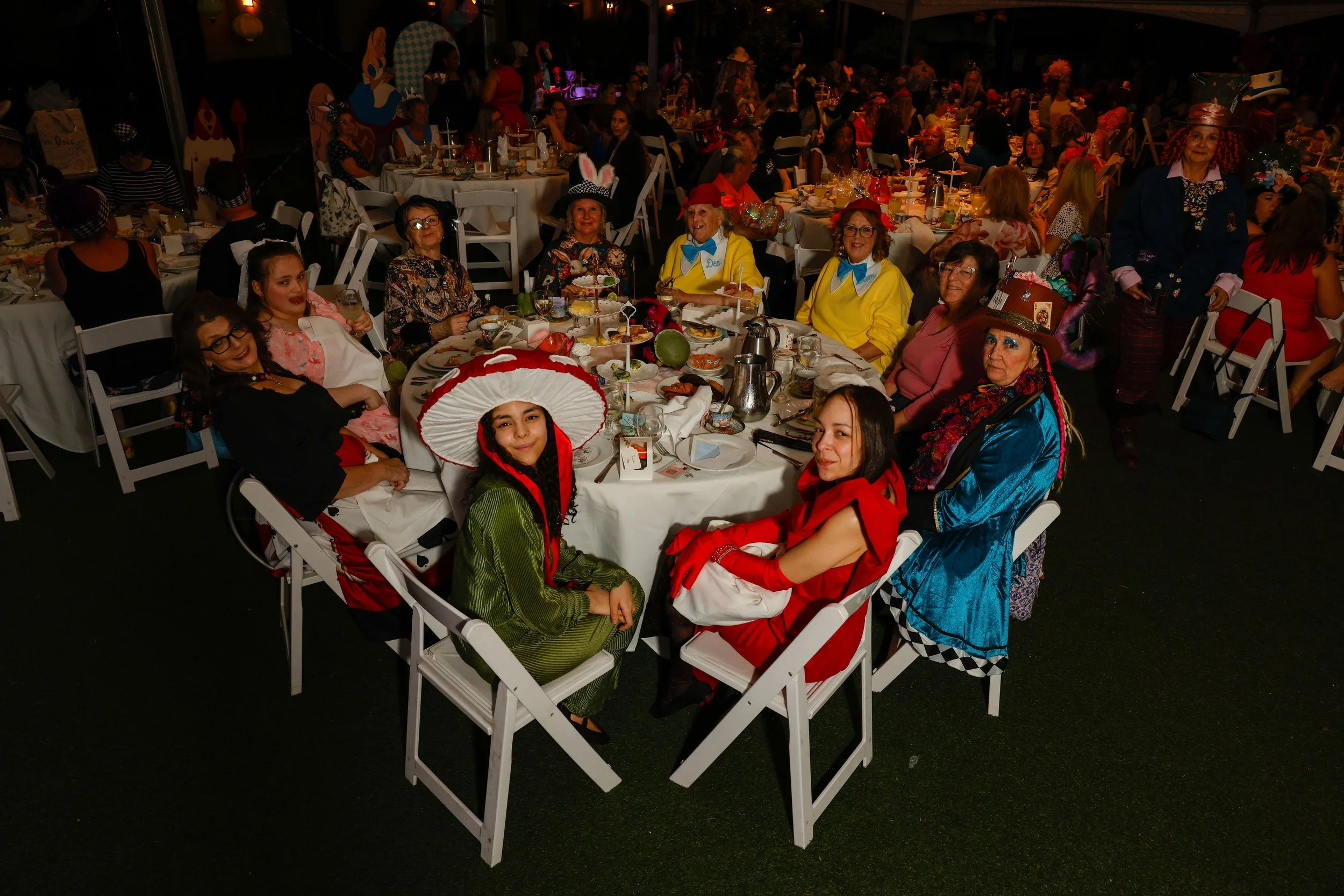 A group of people celebrating at a party, many dressed in costumes including a girl in a mushroom costume and others in colorful outfits and accessories, seated around tables with food and drinks.