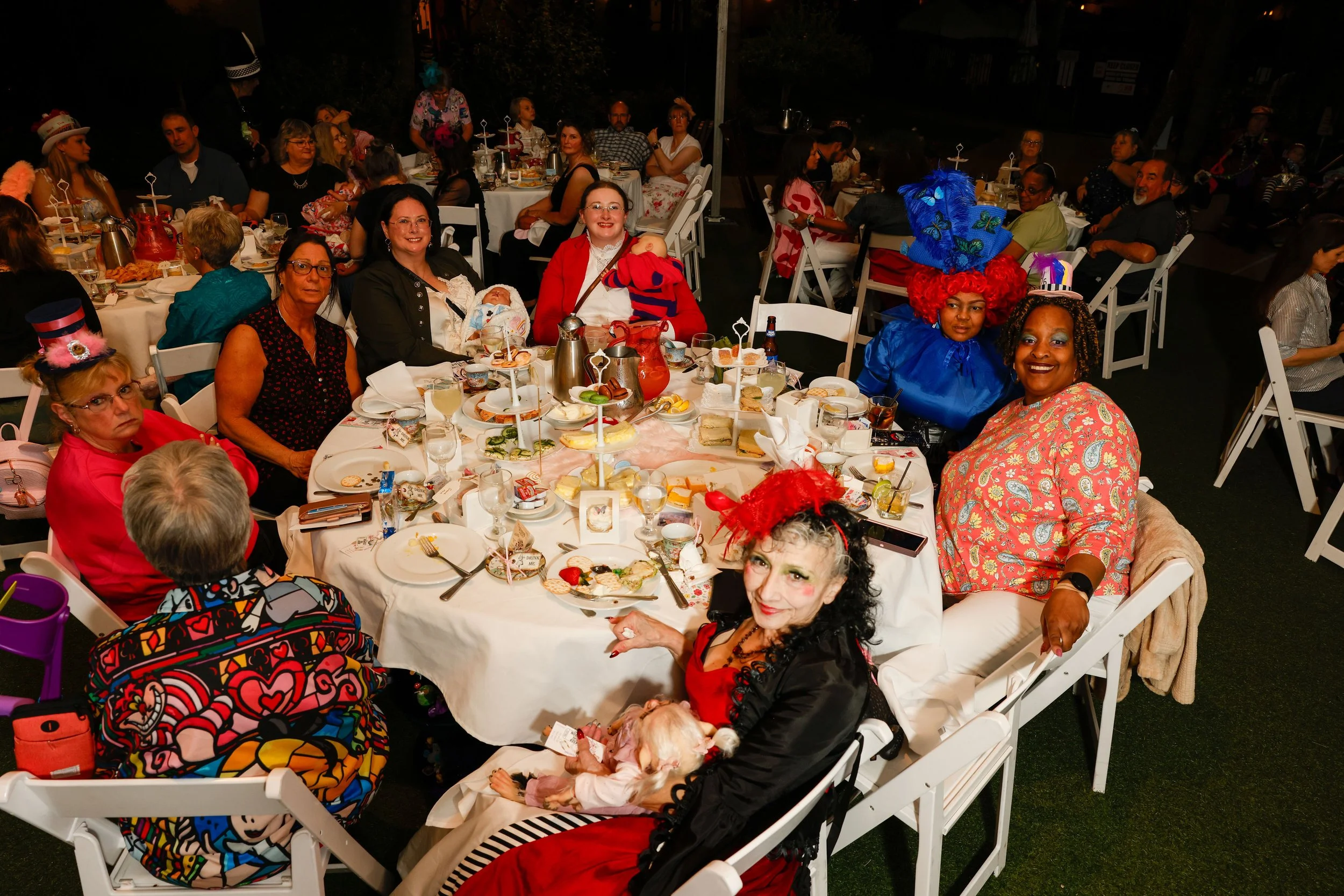 People gathered around a banquet table celebrating a festive occasion. The group includes women dressed in colorful costumes and accessories, including some wearing hats and red clothing. The table is set with plates, glasses, and a variety of food a
