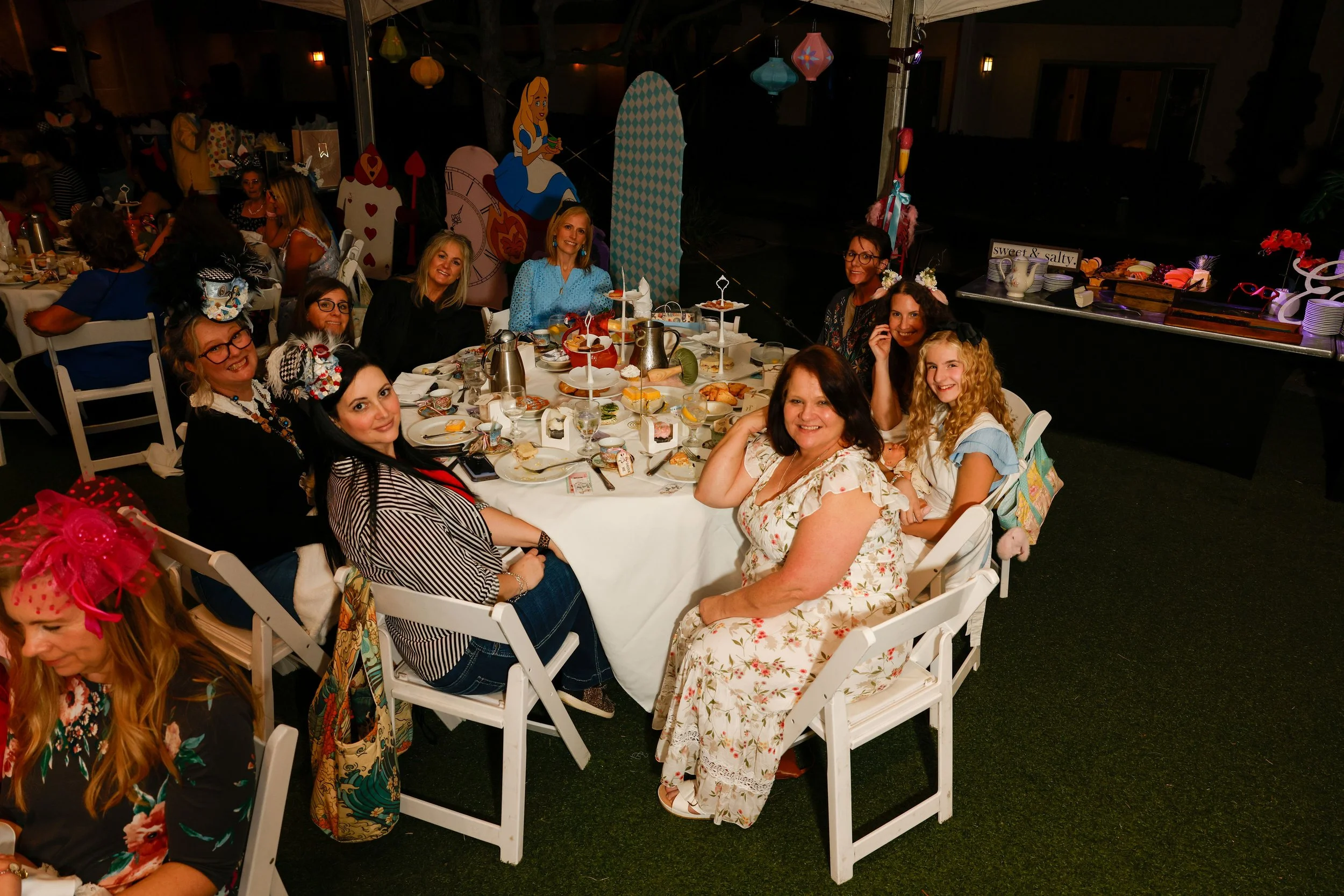 A group of women celebrating together at a party, sitting around a table with food and drinks. The background features Alice in Wonderland-themed decorations, including characters and large playing card motifs.
