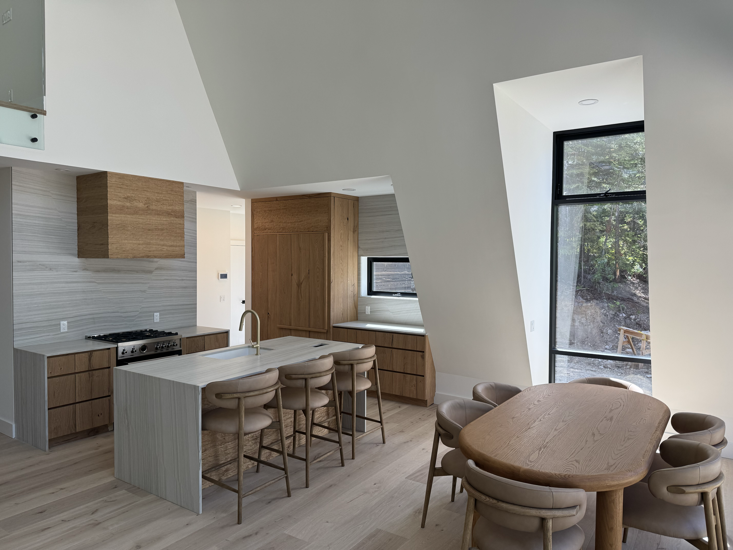 Modern kitchen and dining area with light wood cabinets, a marble-like backsplash, a kitchen island with four chairs, and a dining table with six chairs, featuring large windows with views of trees outside.