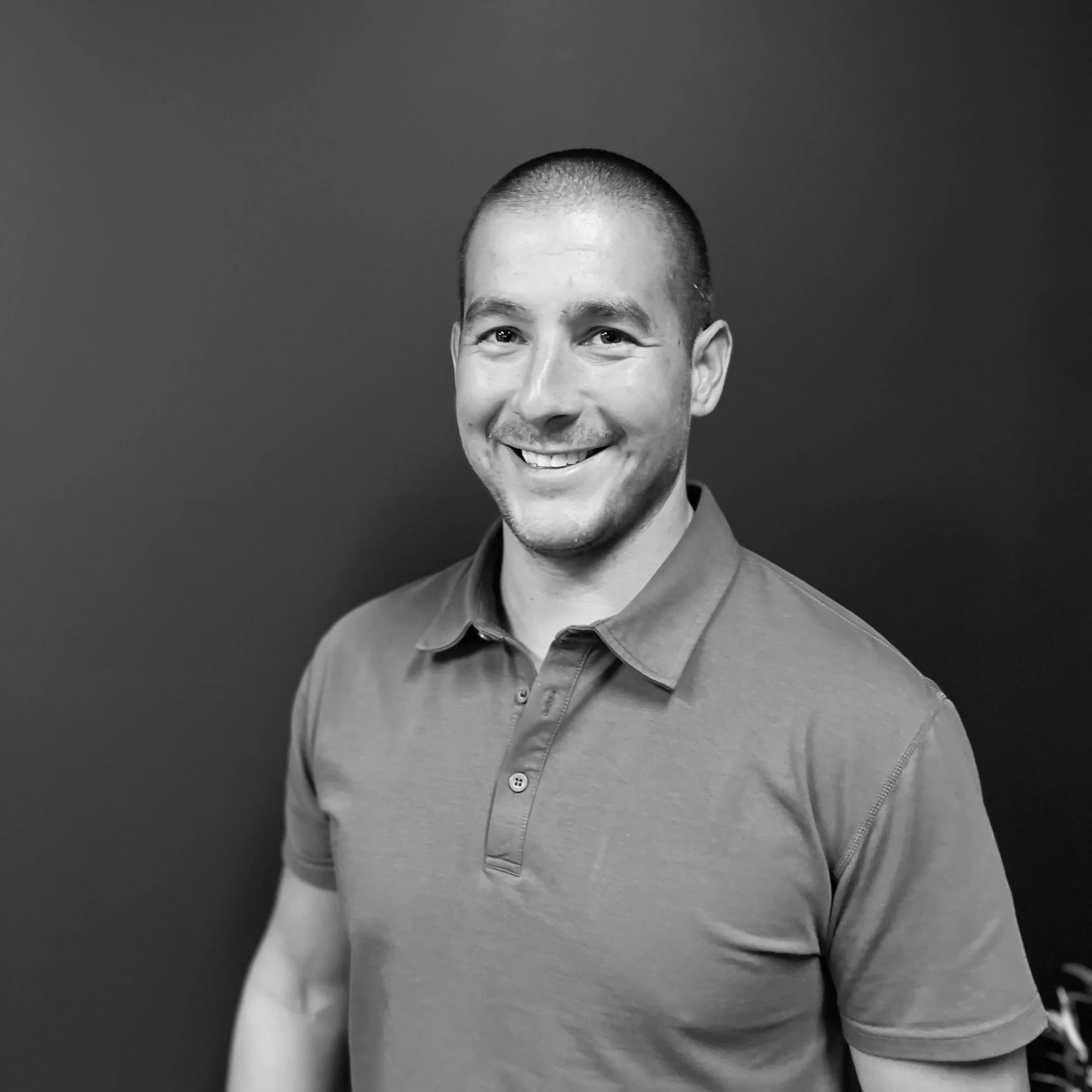 A smiling man with short hair wearing sunglasses and a checkered shirt outdoors in a rocky, mountainous area.