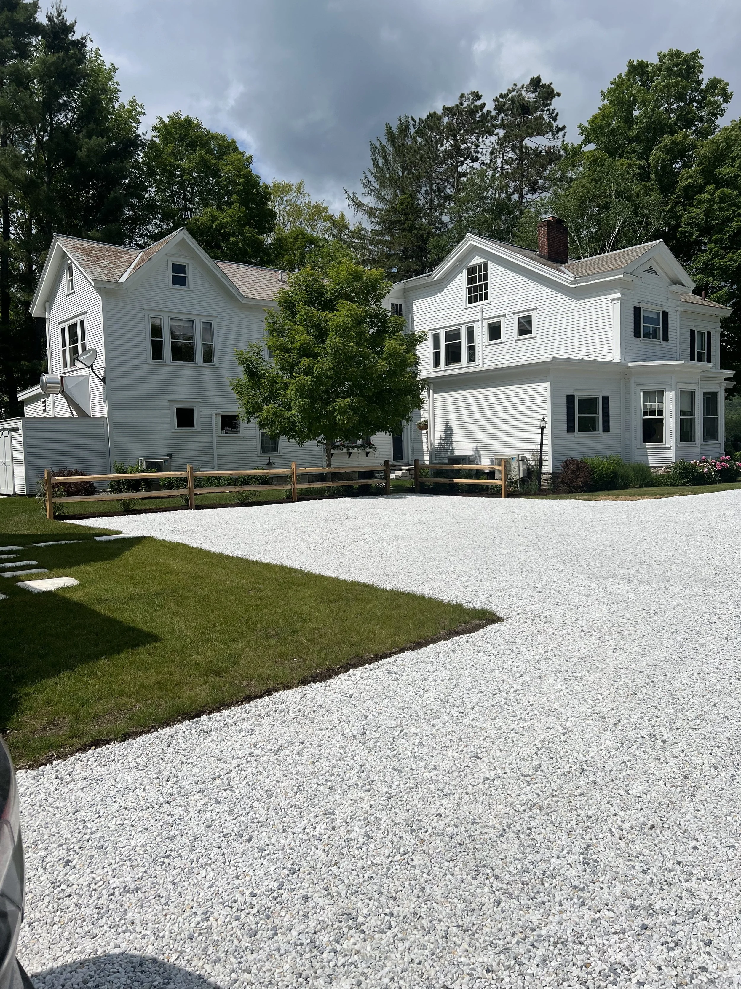 Two large white houses with multiple windows and black shutters, surrounded by green trees, on a sunny day with cloudy sky, gravel driveway in front, small fence and garden on the side.