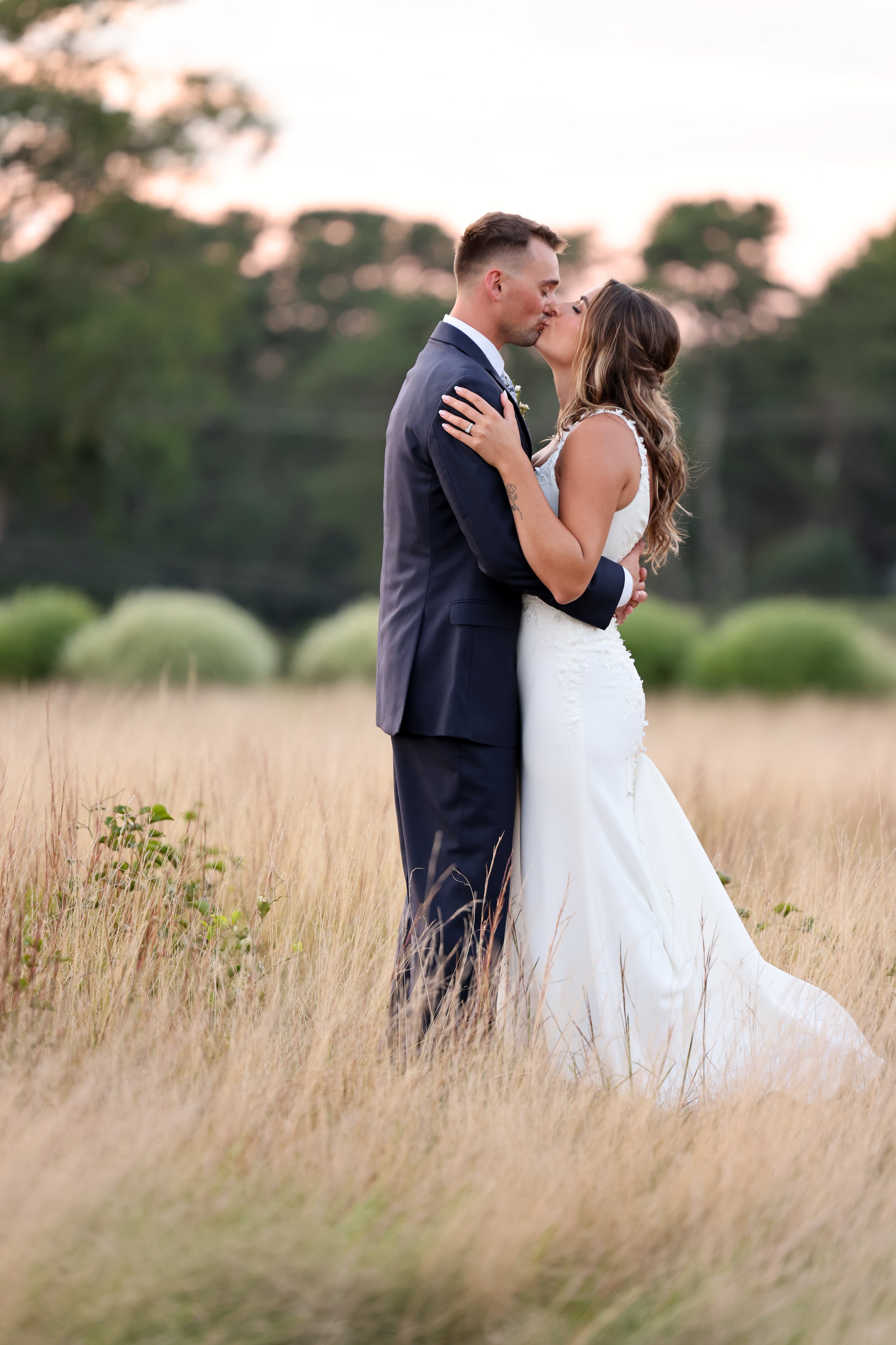 A newlywed couple kissing in a field with tall grass at sunset, the groom in a dark suit and the bride in a white wedding dress.
