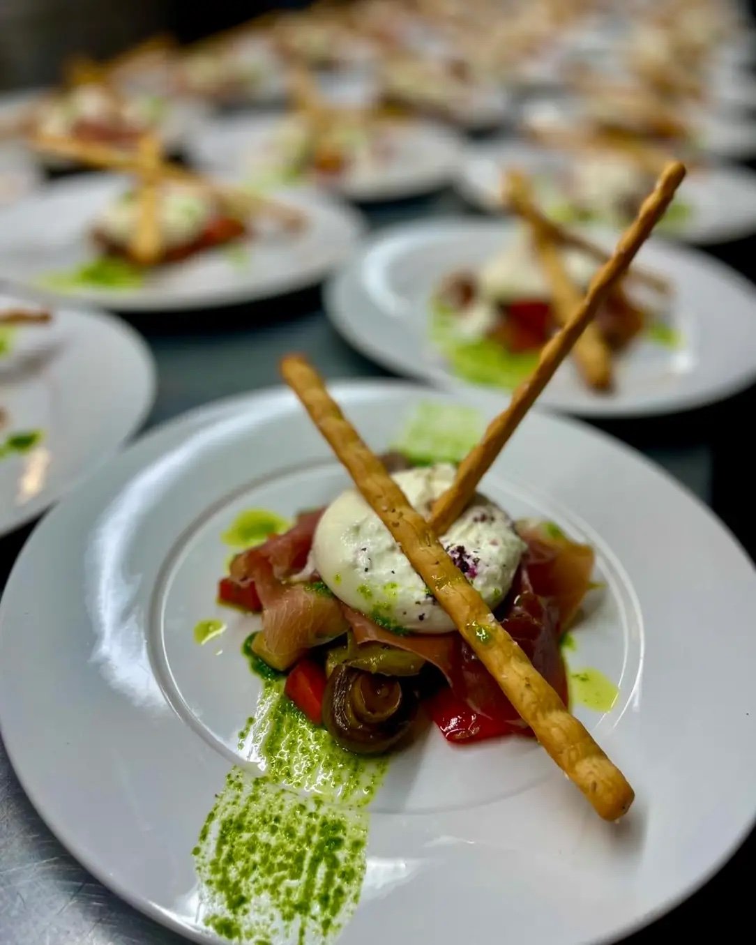 Gourmet-Vorspeise auf einem weißen Teller, bestehend aus einem Käse oder einer Creme, Schinken oder Lachs, Tomaten, einer Zwiebel und Grissini-Stangen, garniert mit einer grünen Sauce. Im Hintergrund sind ähnliche Teller unscharf zu sehen.