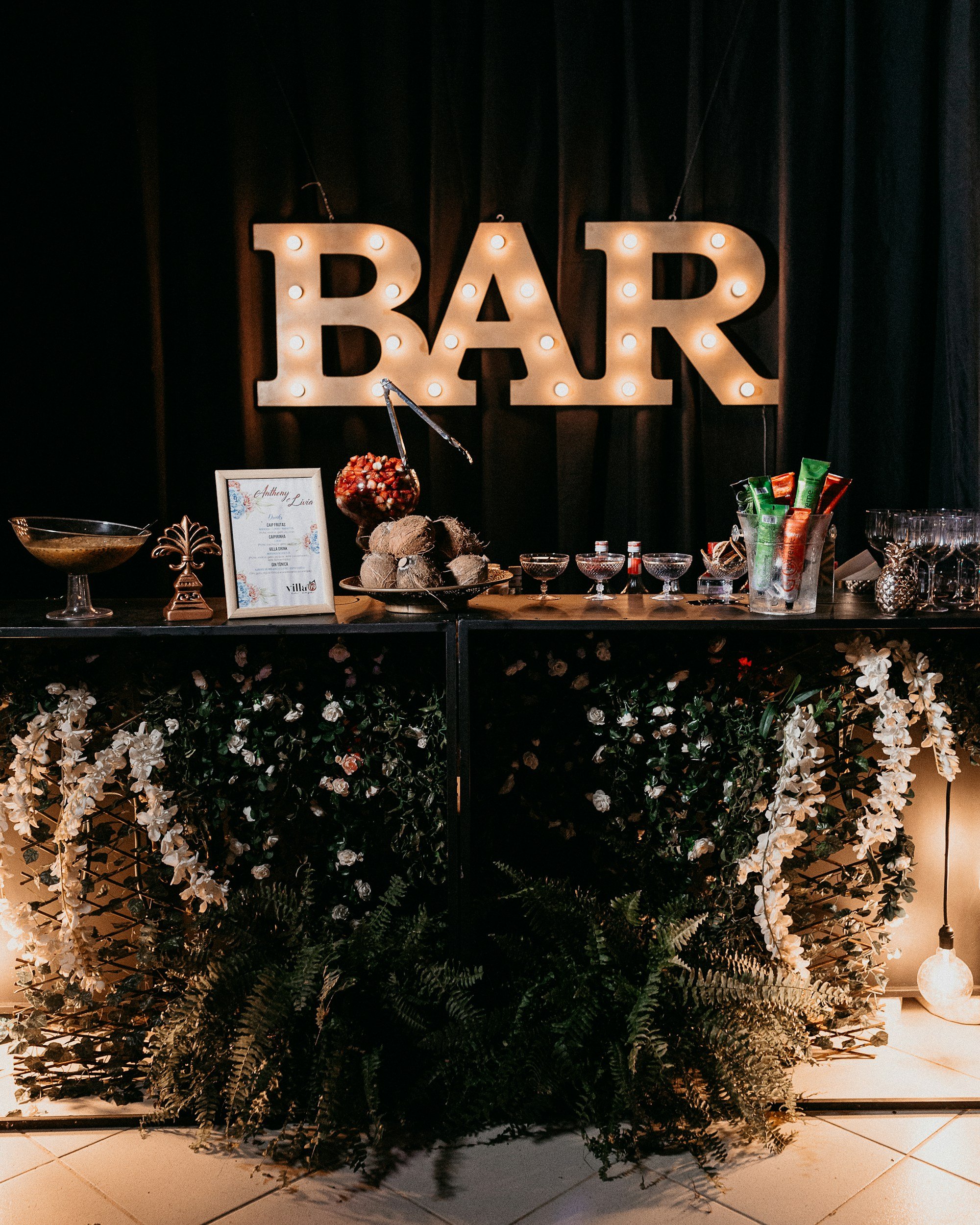 Eine Bar mit beleuchtetem "BAR"-Schriftzug, dekoriert mit Pflanzen und Blumenarrangements. Auf der Theke stehen Gläser, Flaschen und verschiedene Barutensilien. Im Vordergrund sind Farnpflanzen und sanfte Beleuchtung zu sehen.
