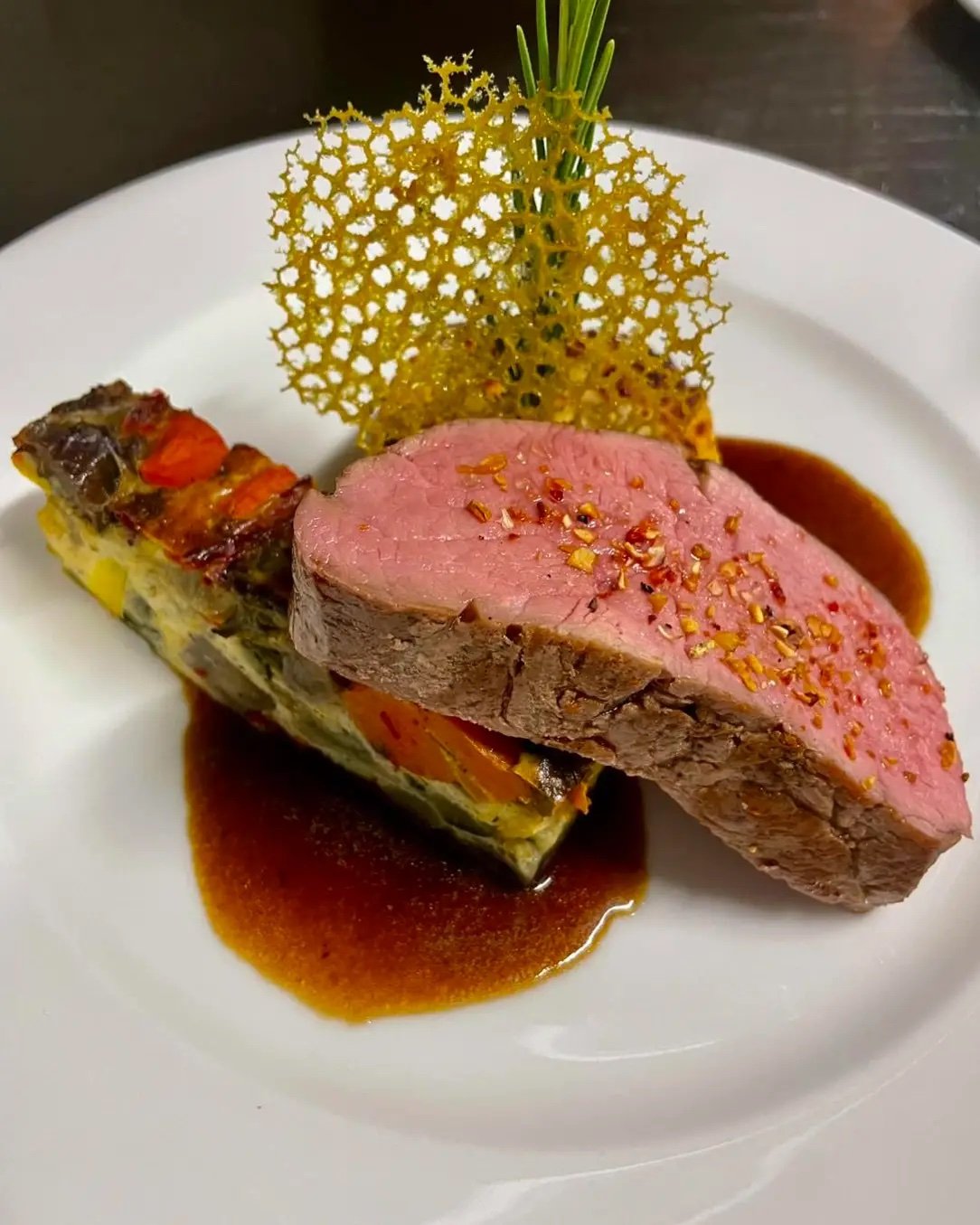 Ein Teller mit einem Stück rosa gebratenem Fleisch, einer Gemüsequiche, einer dunklen Sauce und einer dekorativen, goldenen Netzkruste, garniert mit einem grünen Zweig.